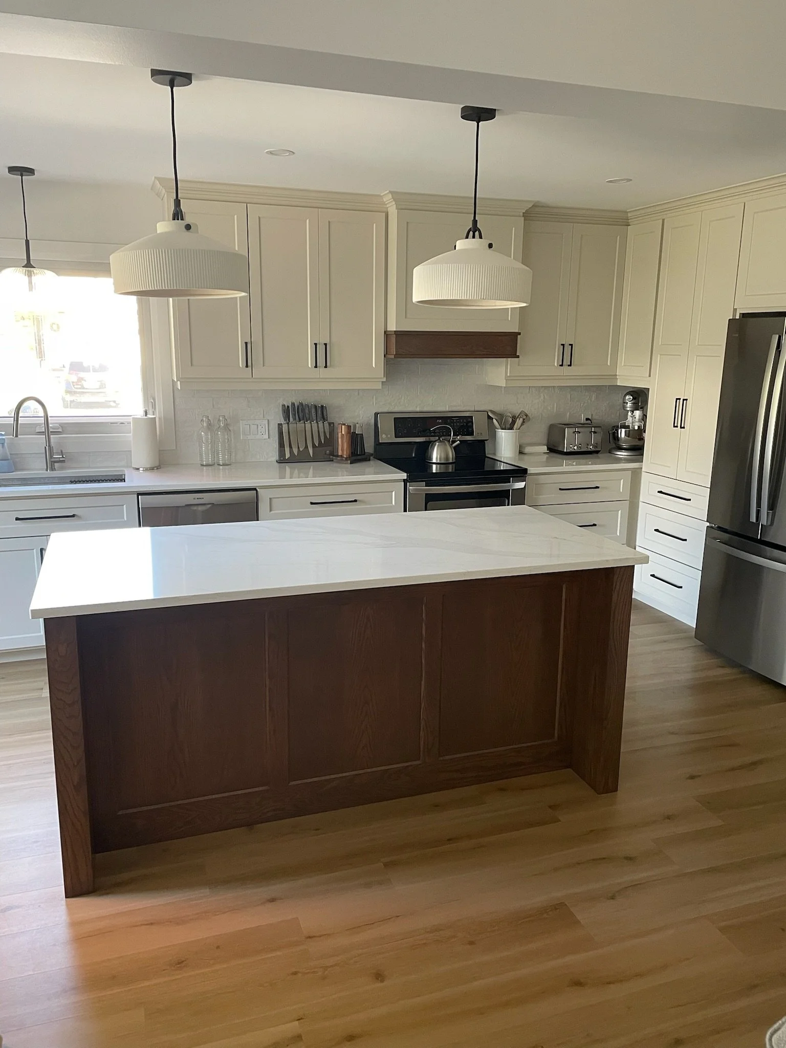 A modern kitchen with white cabinets, stainless steel appliances, a white quartz countertop, and pendant lights hanging above the island. There is a window letting in natural light and various kitchen utensils on the counter.