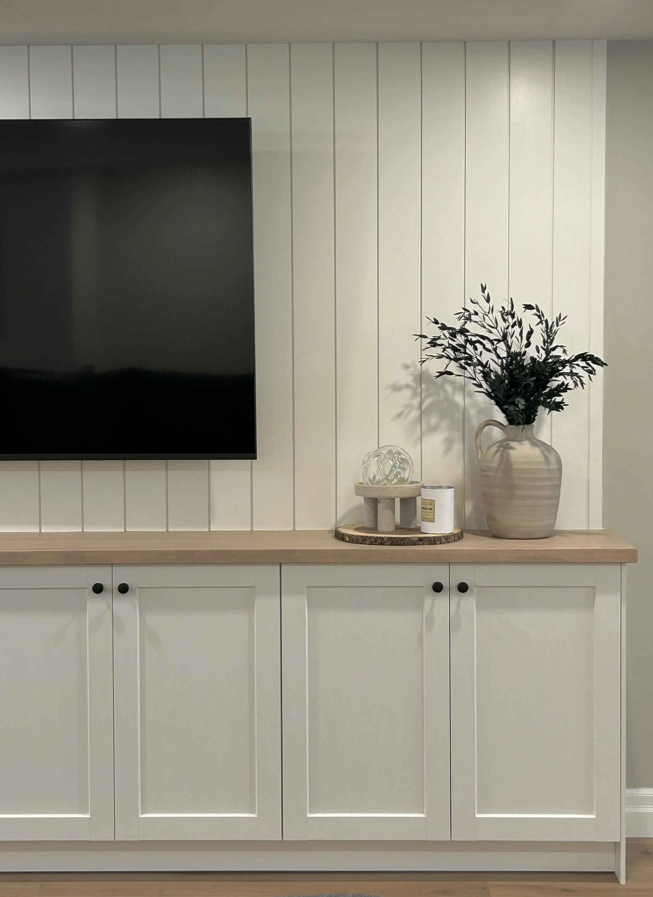 A white wall with vertical shiplap paneling, mounted on a white cabinet with black knobs and wood counter. On top of the cabinet, there is a round wooden tray with decorative glass objects, a beige vase with leafy branches.