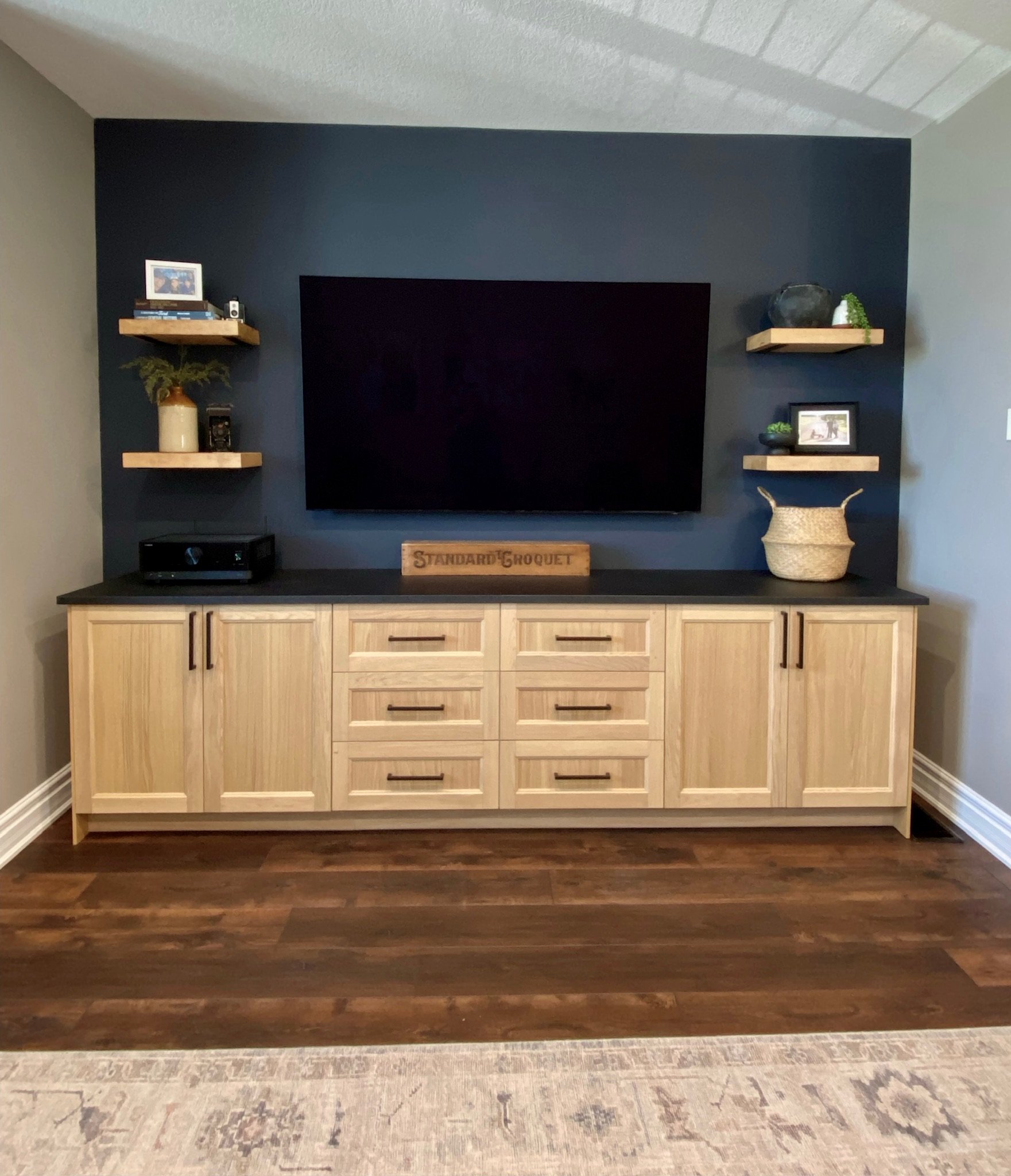 Living room entertainment center with a large black TV mounted on a dark accent wall. There are wooden shelves with decorative items and picture frames on either side of the TV. A wooden cabinet with drawers and doors is positioned below the TV, and 