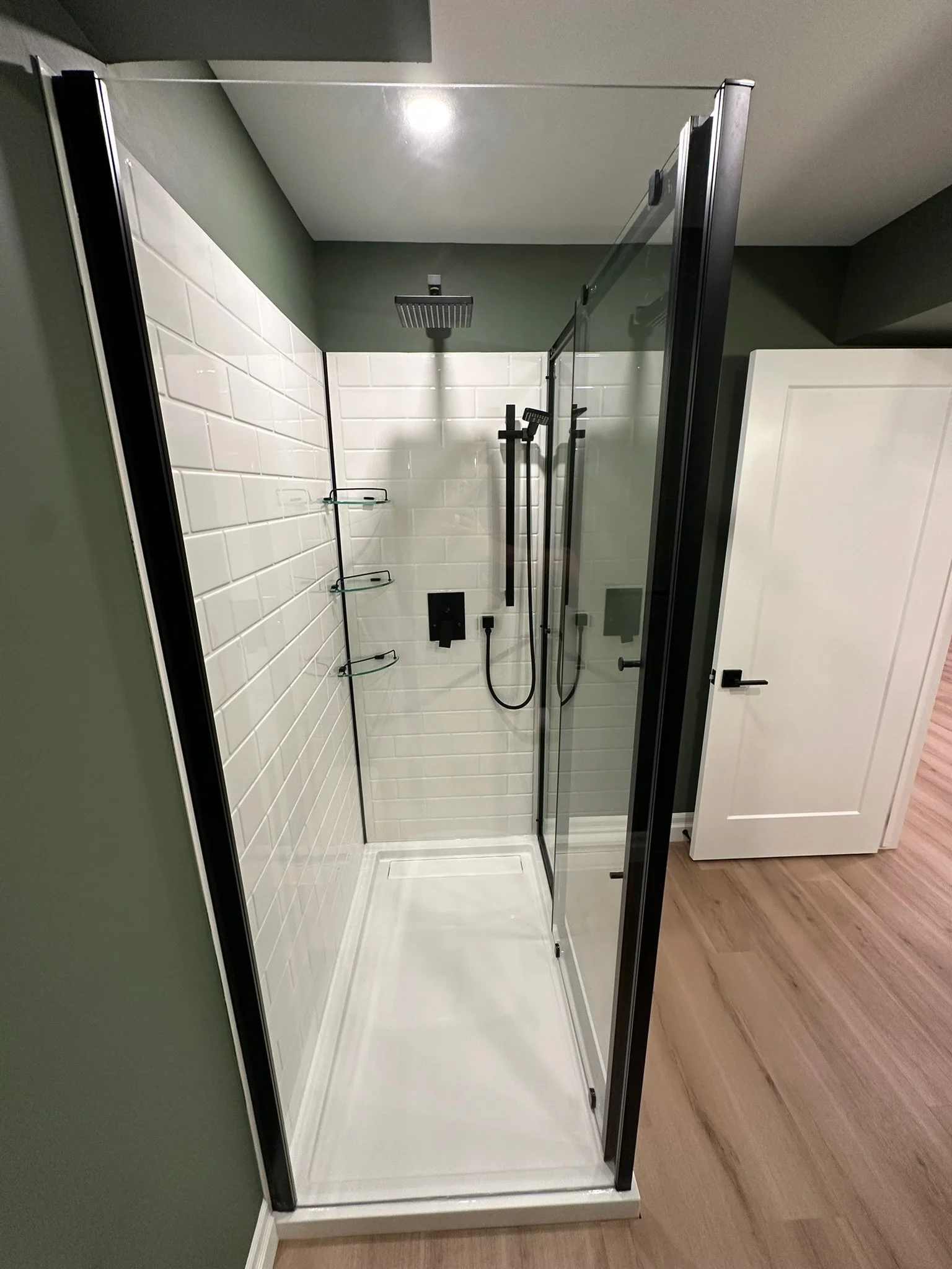 A modern glass shower enclosure with black frame and white tiled walls, mounted showerhead, and a black shower panel, in a bathroom with green and white walls and wood flooring.