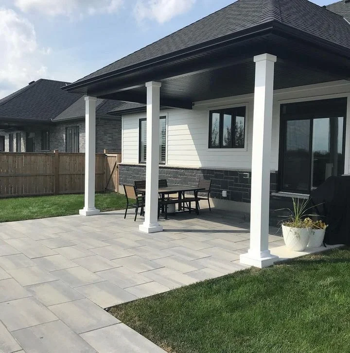 Backyard patio with a covered seating area, white PVP columns, black roof, modern furniture, potted plant, wooden fence, and neighboring house in the background.