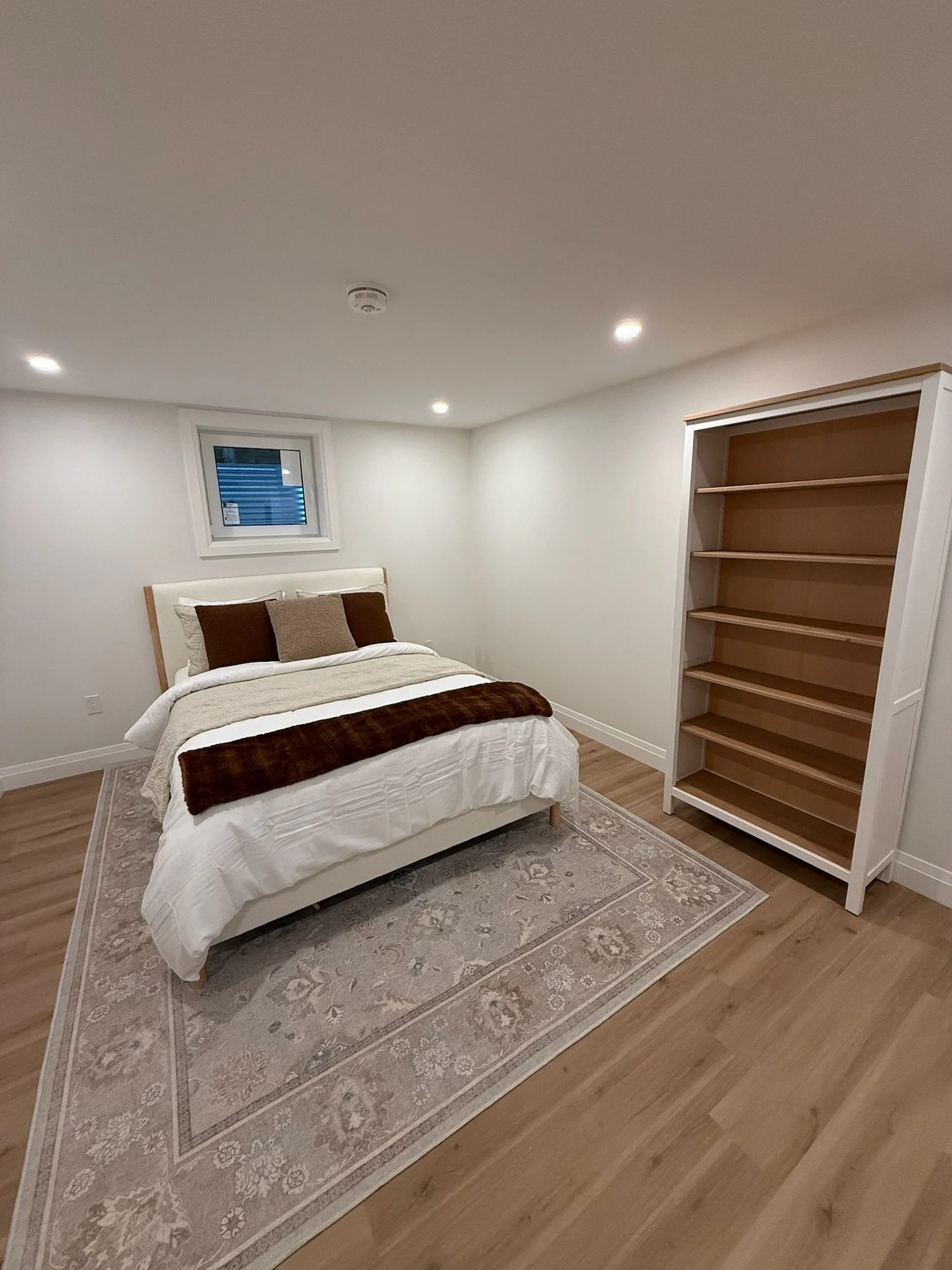 A bedroom with a bed, side pillows, a window, a rug, and a wooden bookshelf.