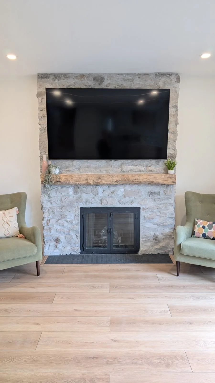 Living room with a stone fireplace, black television mounted above. Two light green armchairs with patterned pillows on either side of the fireplace, and a wooden mantel decorated with small plants and a vase.