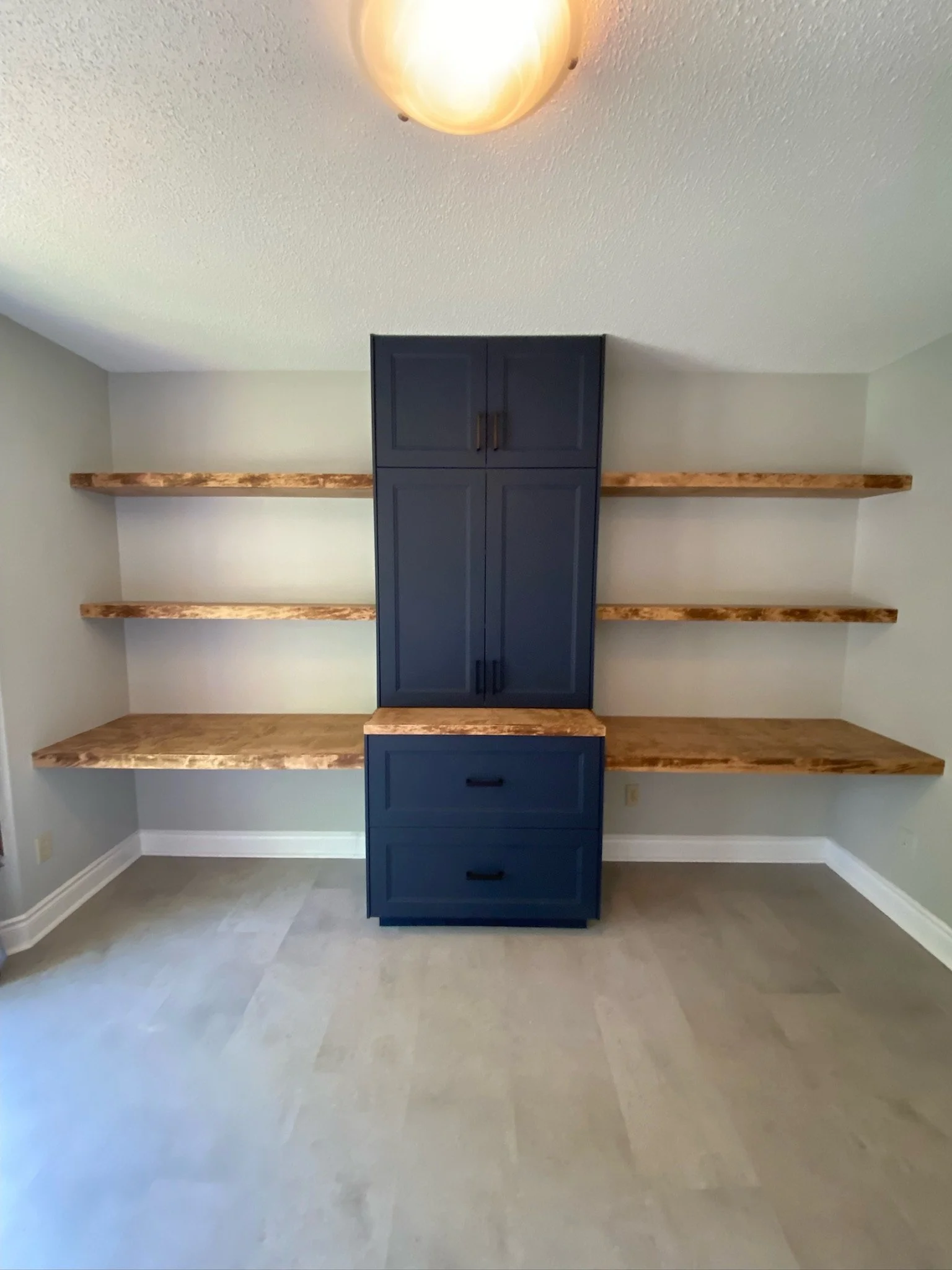 Empty room with a blue built-in cabinet with four drawers in the center, three wooden shelves on the sides, beige walls, a textured white ceiling with a ceiling lights, and beige tile flooring.
