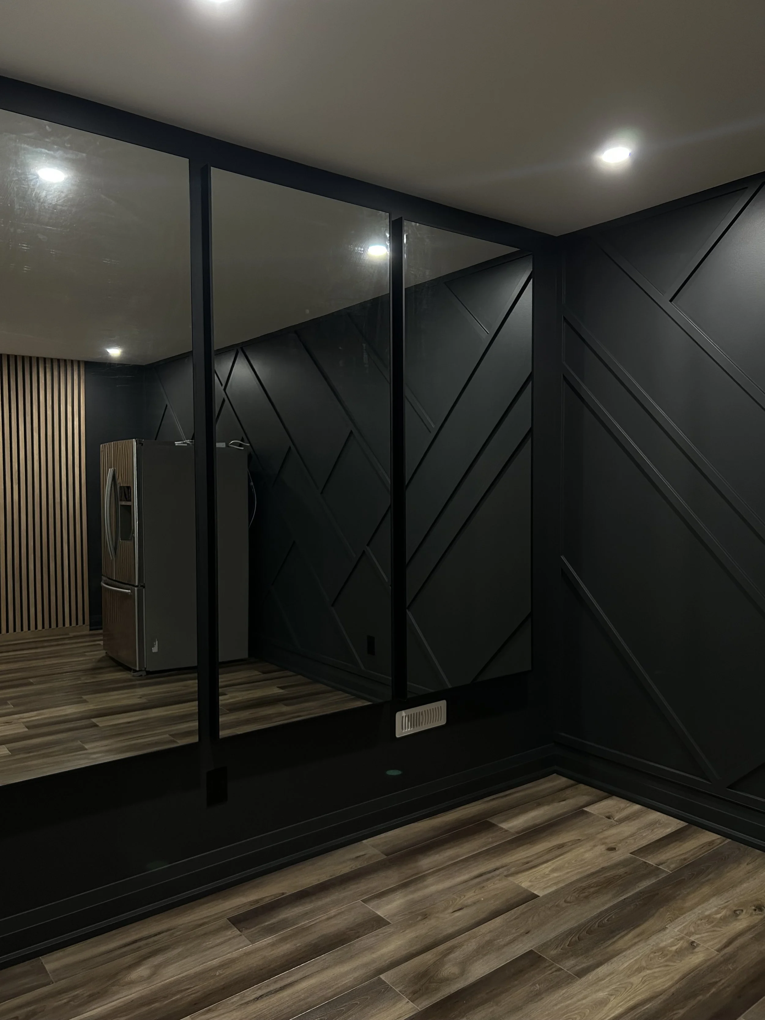 A modern kitchen corner featuring a black geometric wall design, a stainless steel refrigerator, a wood-paneled wall, and a large mirror reflecting ceiling lights, with a wooden floor.