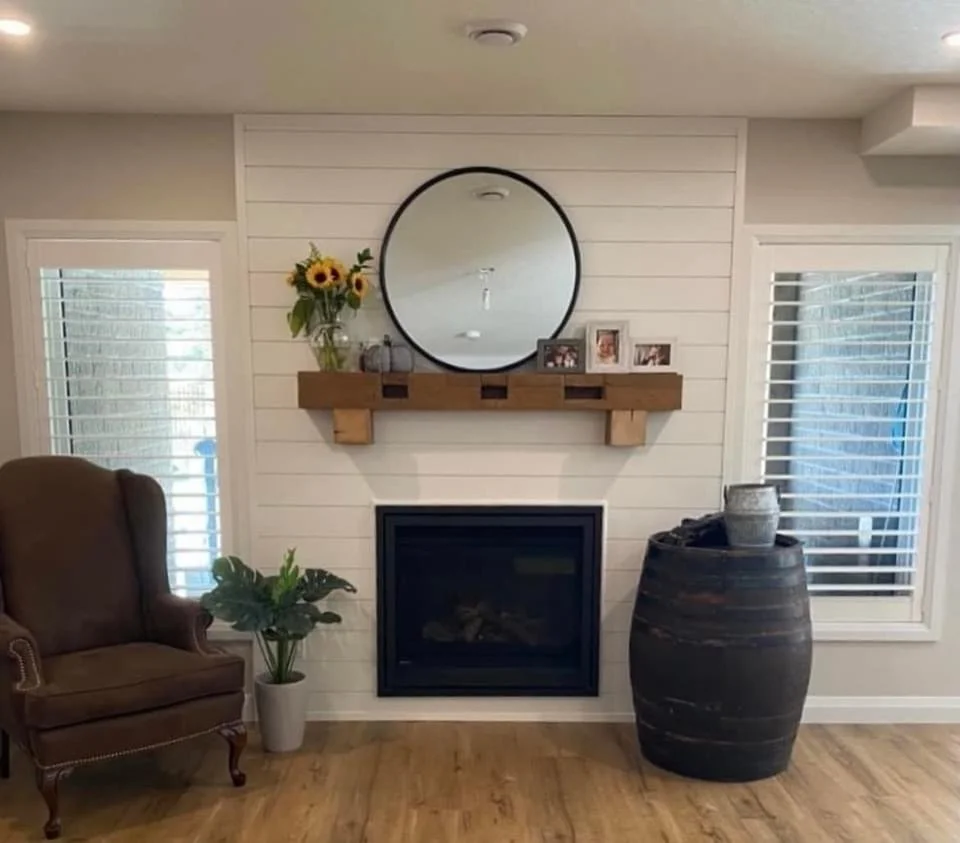 Living room fireplace with a round mirror above a wooden mantel, framed photos, sunflower arrangement, a potted plant, a large decorative vase, and nearby windows with white blinds.