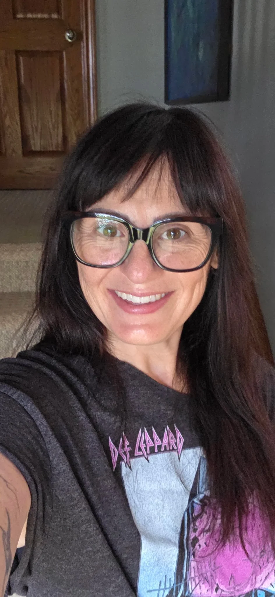Smiling woman with long dark hair, glasses, and t-shirt taking selfie indoors near a wooden staircase and framed art on the wall.