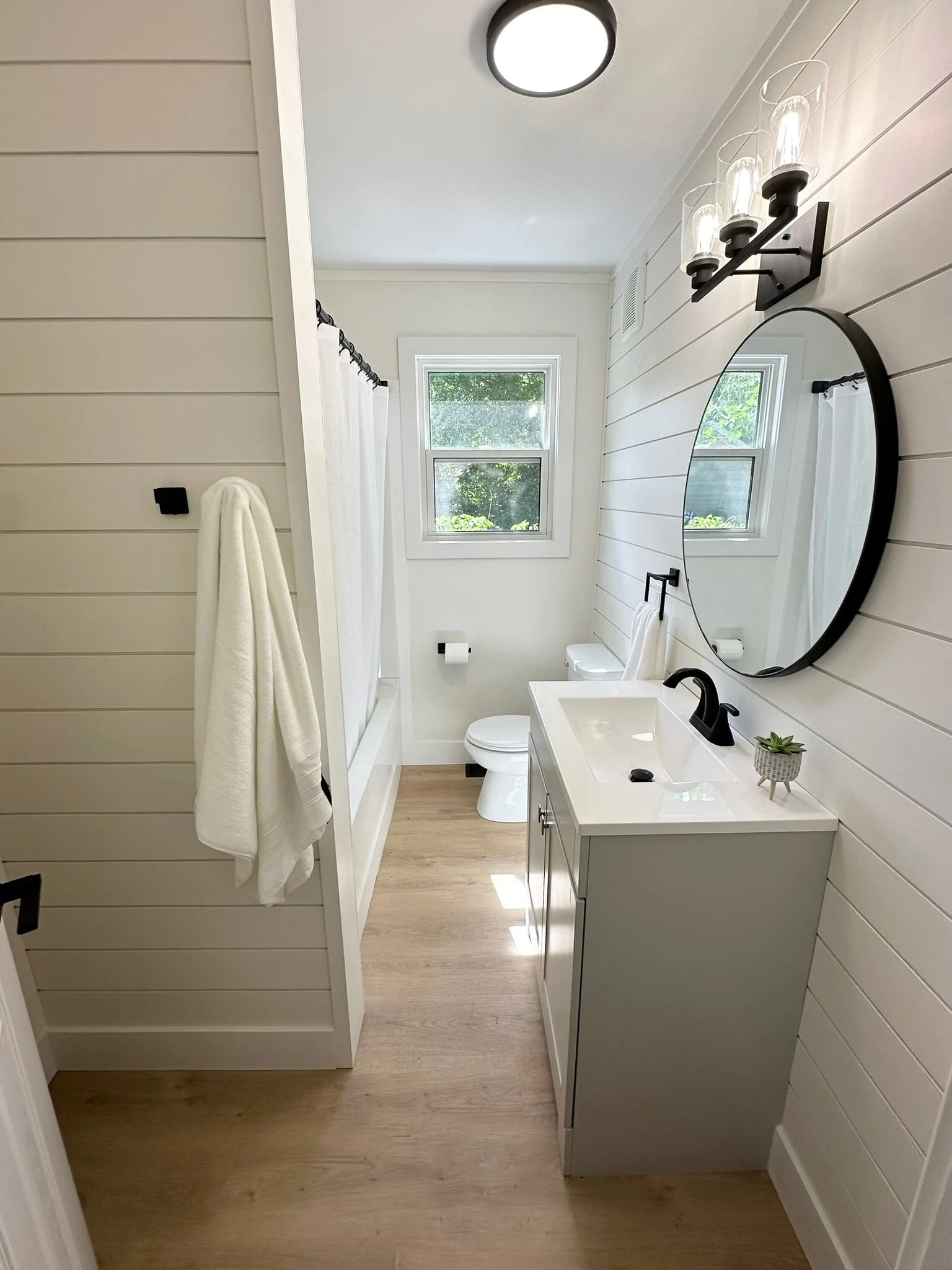 A modern bathroom with white shiplap walls, a large window, a white vanity with a black faucet, a round mirror, a three-light fixture, a toilet, and a shower with a curtain. There is a towel hanging on a hook and a small potted succulent on the vanit
