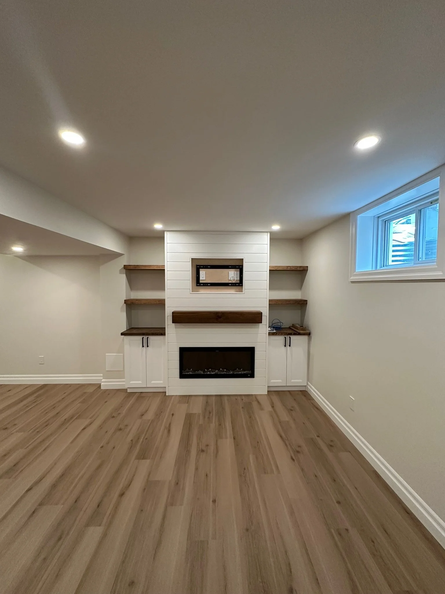 Empty living room with hardwood floors, white walls, a central fireplace with a white shiplap surround, built-in wooden shelves on either side, a window on the right side, and recessed ceiling lights.