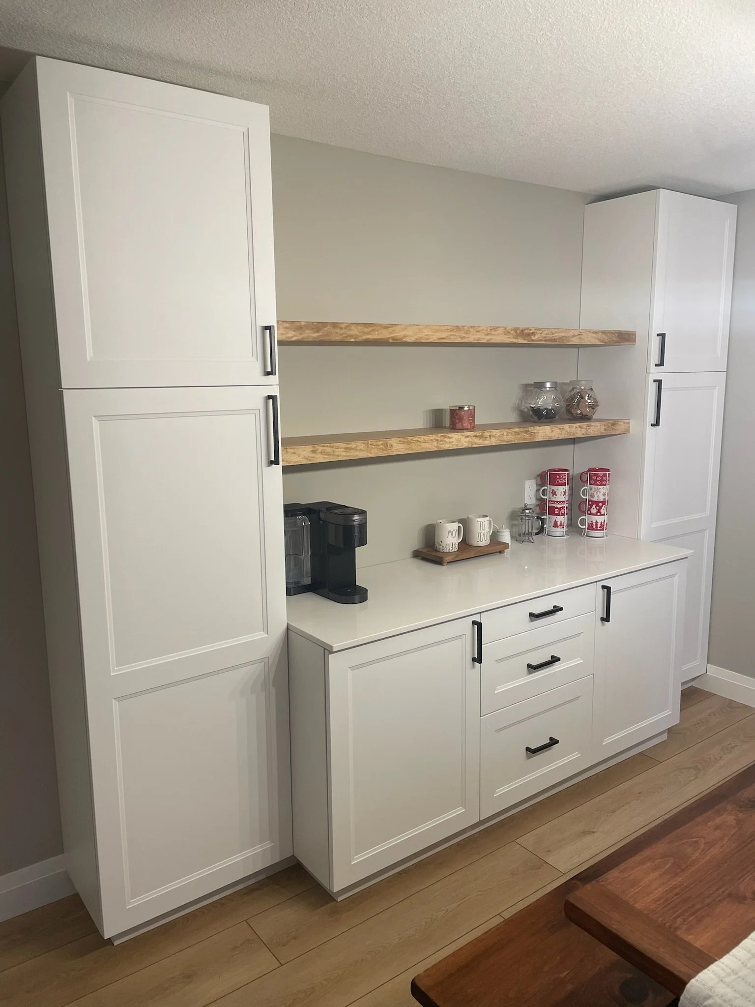 A kitchen with white cabinets, wooden shelves, and various kitchen items including mugs, jars, and a coffee maker, with hardwood flooring.