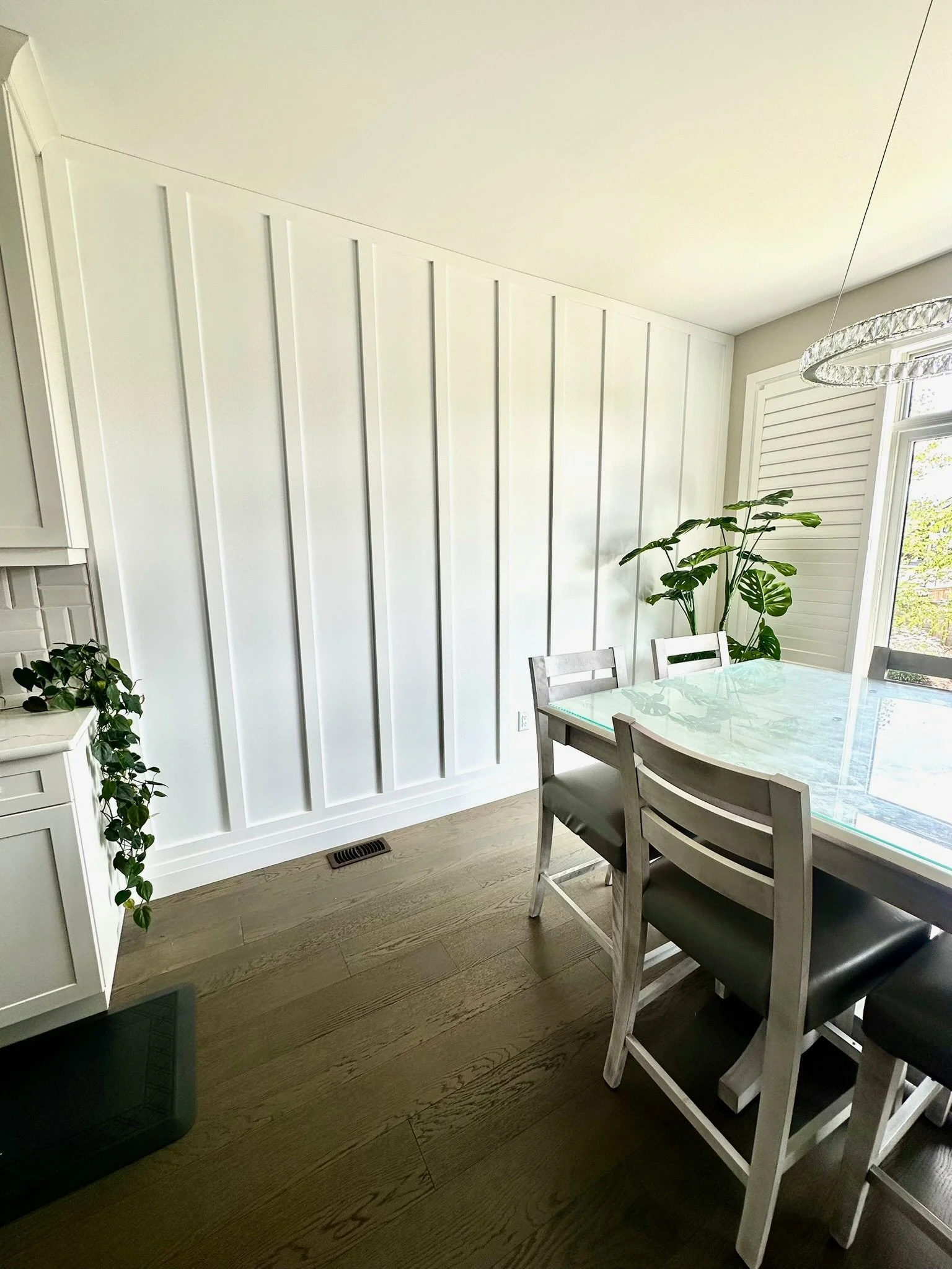 Dining area with a glass top table, white chairs, large window with shutters, potted plants, hardwood floor, and a modern light fixture. The accent wall add a subtle but textural element to this space. 