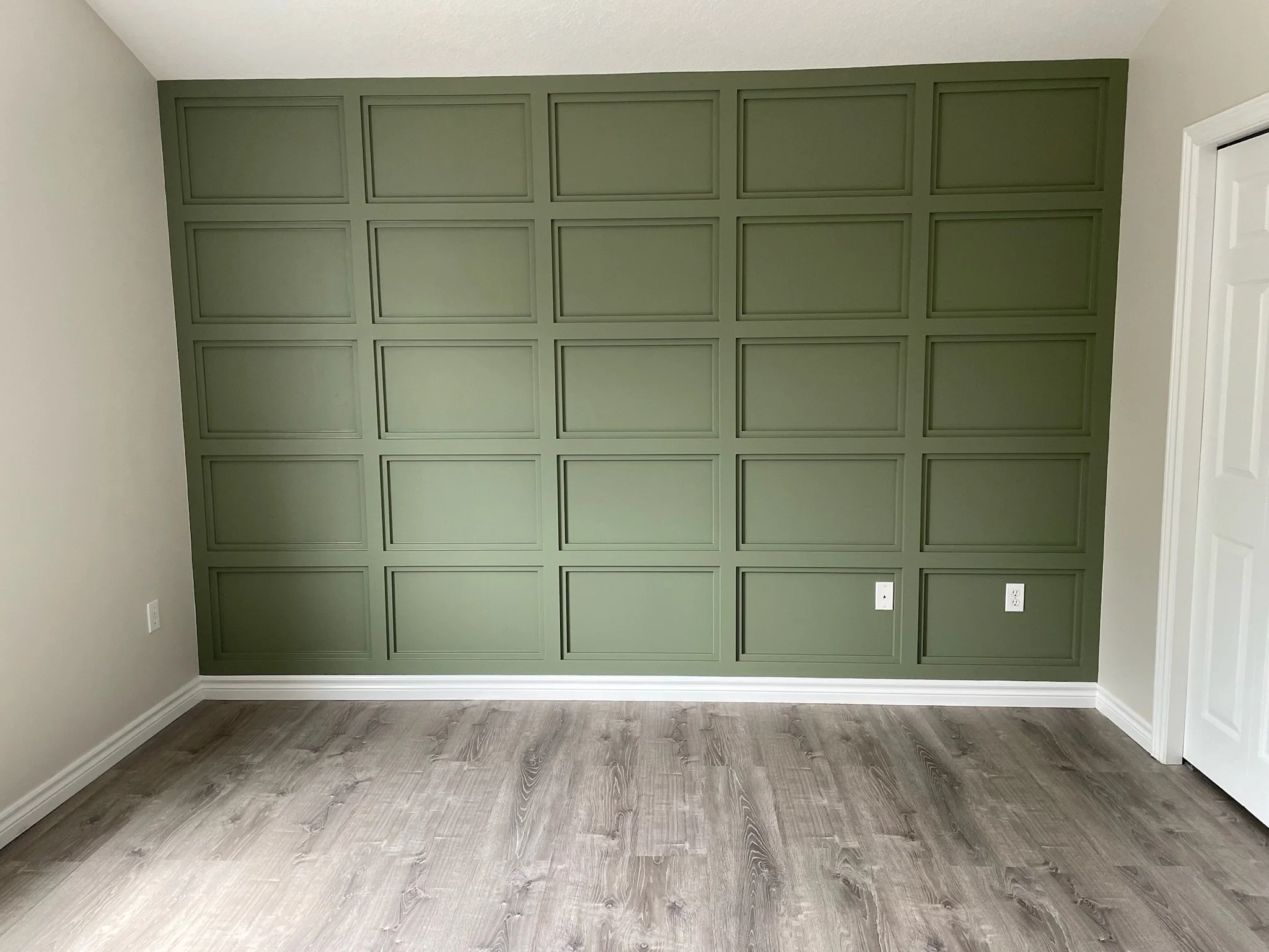 Interior of a room with a green wall featuring a decorative wall panel design, white baseboards, and light wood flooring.