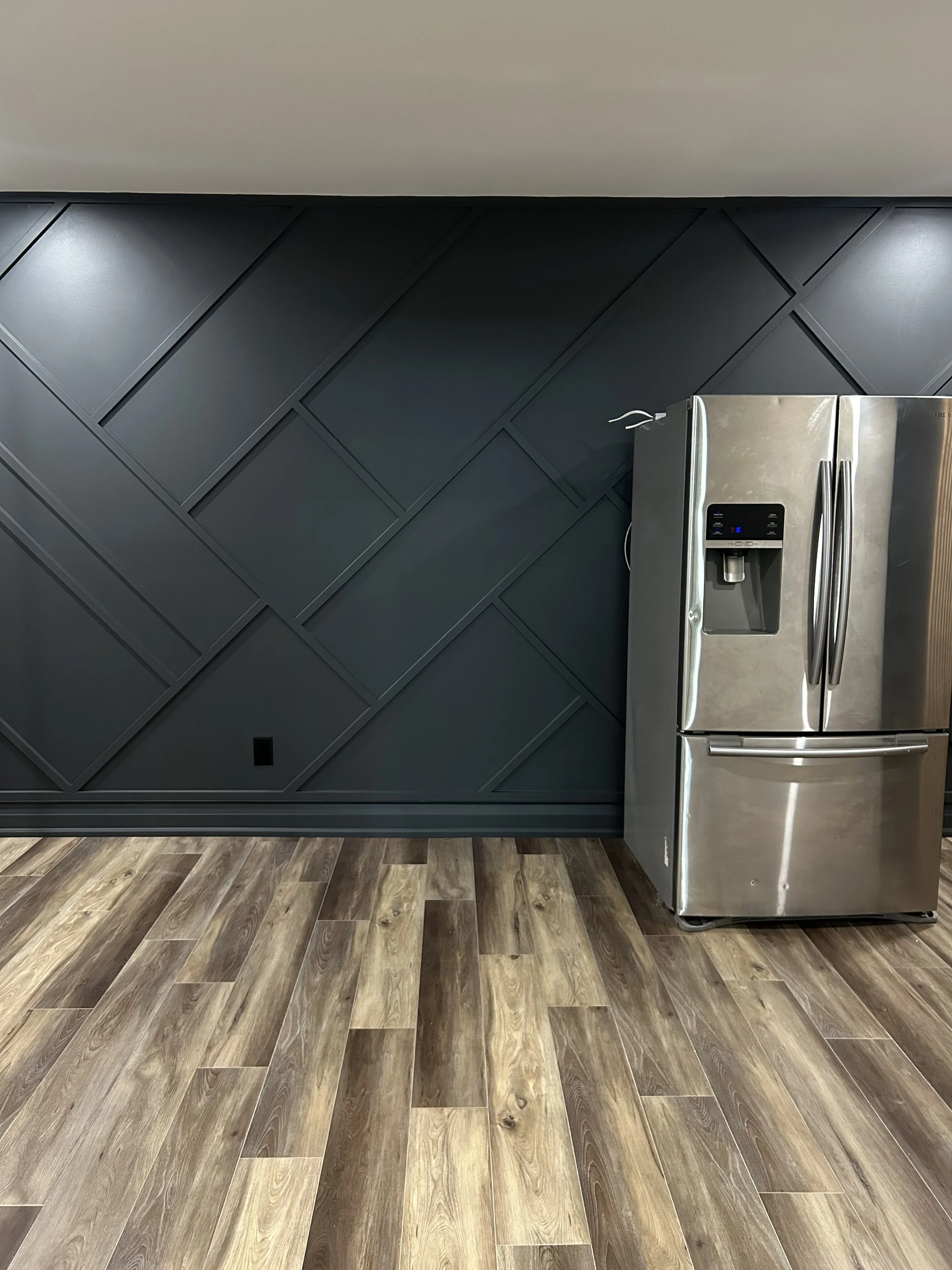 A stainless steel refrigerator with a water and ice dispenser in a modern kitchen, dark accent wall with a geometric pattern, and wood-colored floor tiles.