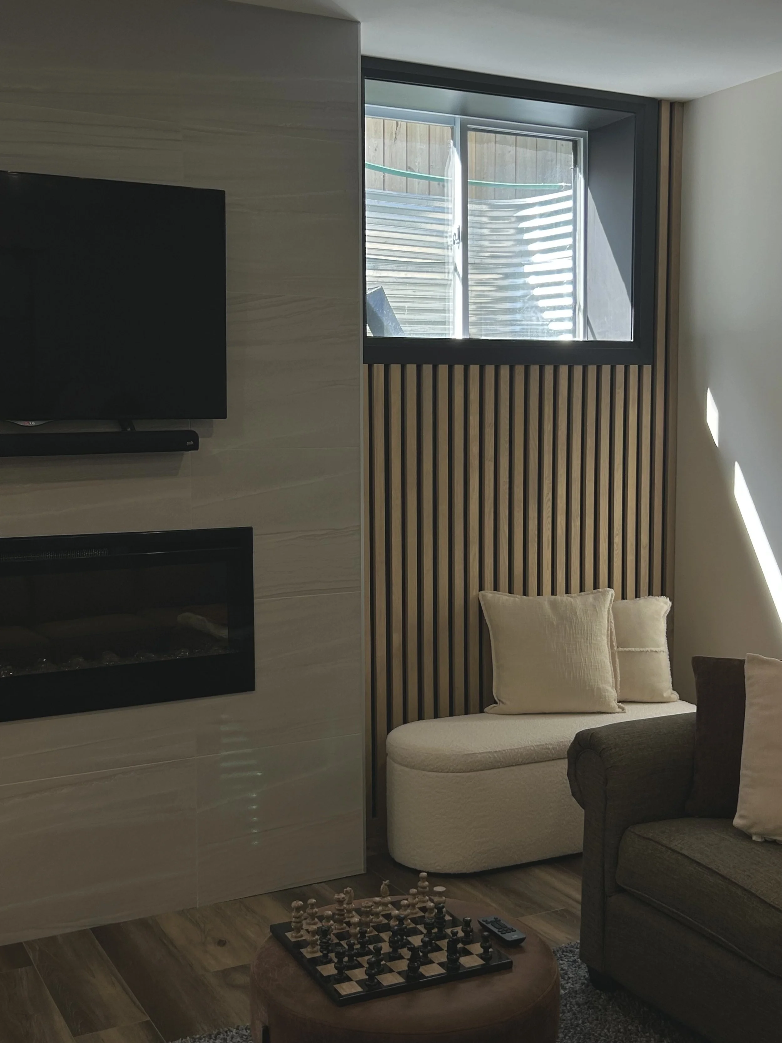 A cozy living room with a beige sofa, a dark armchair, and a chessboard on a round ottoman. There is a large window with wooden slats and a partial fireplace or cabinet on the left side. The floor is wooden, and sunlight is coming through the window.
