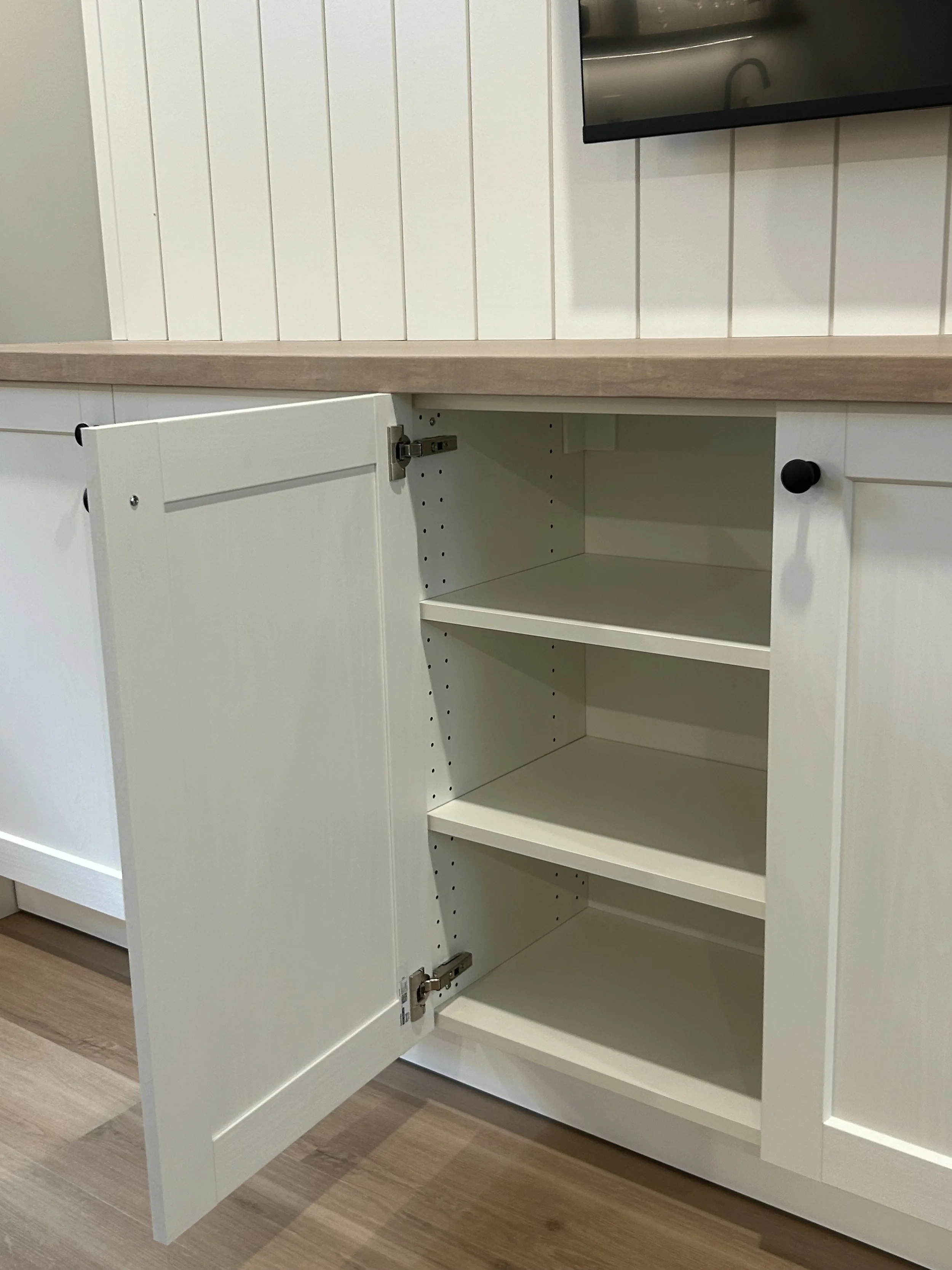 Empty white kitchen cabinet with open door, showing shelves inside, in a room with wood floor and shiplap wall.