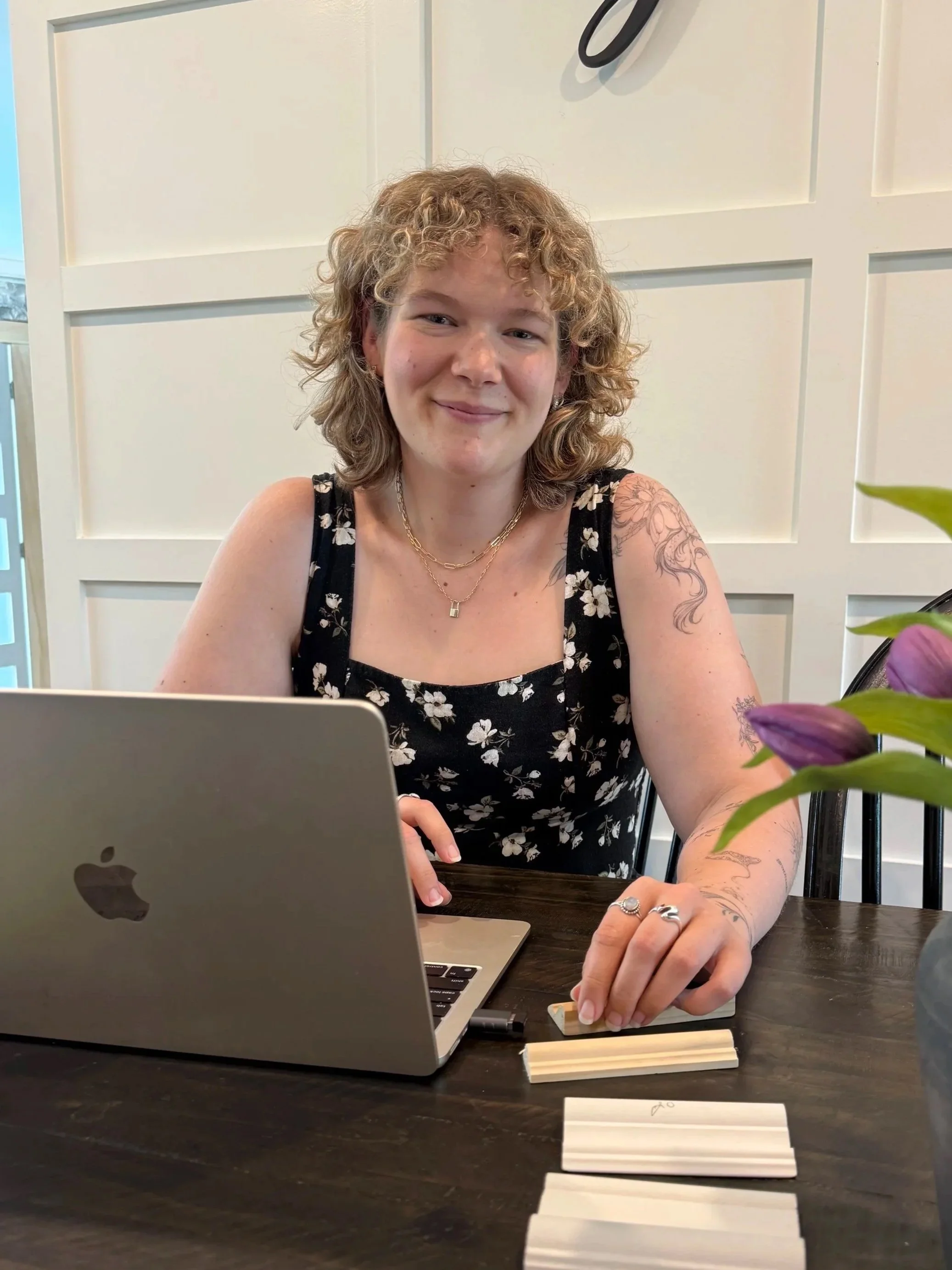 A woman with curly hair and tattoos on her arm sitting at a dark wooden table, smiling slightly, with a silver MacBook in front of her, and purple tulips in the foreground.