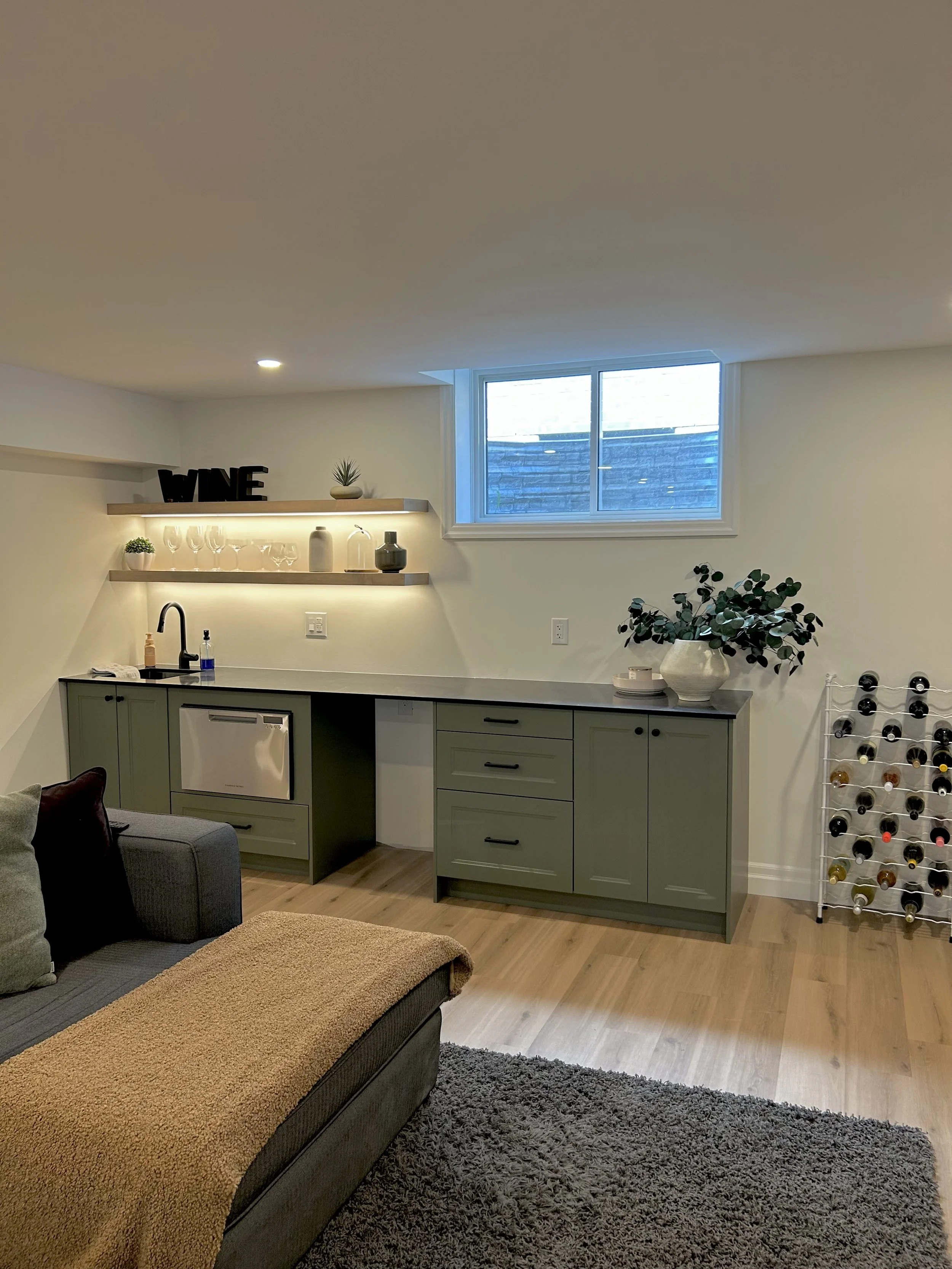 A living room with a kitchenette area featuring green cabinets, a black countertop, and a small sink. Open shelves above hold wine glasses, vases, and decorative items, including a small potted plant and the word 'WINE' in black letters. A window wit
