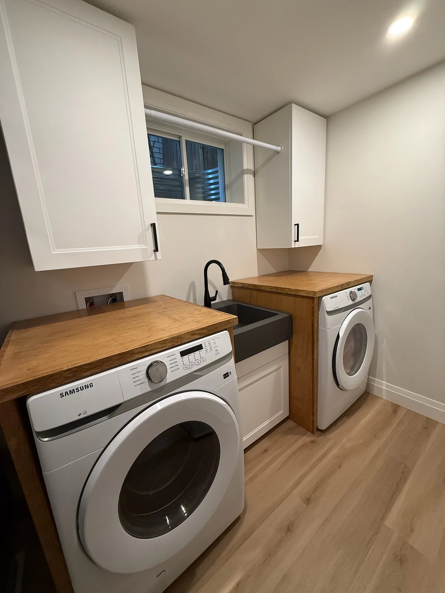 A laundry room with two white Samsung front-loading washing machines side by side beneath a wooden countertop. There are white cabinets above with black handles, and a black undermount sink with a black faucet in between. A window is above the counte