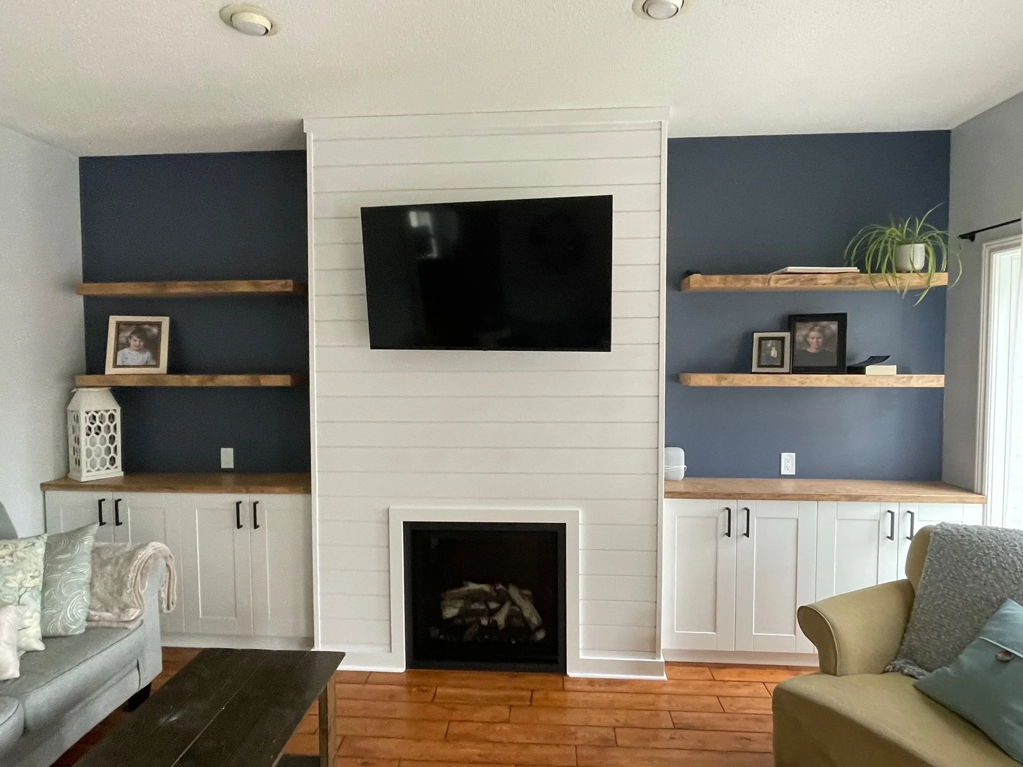 Living room with built-in white cabinetry, a black fireplace, floating wood shelves decorated with framed photos and a potted plant, and a wall-mounted TV on a dark blue accent wall.
