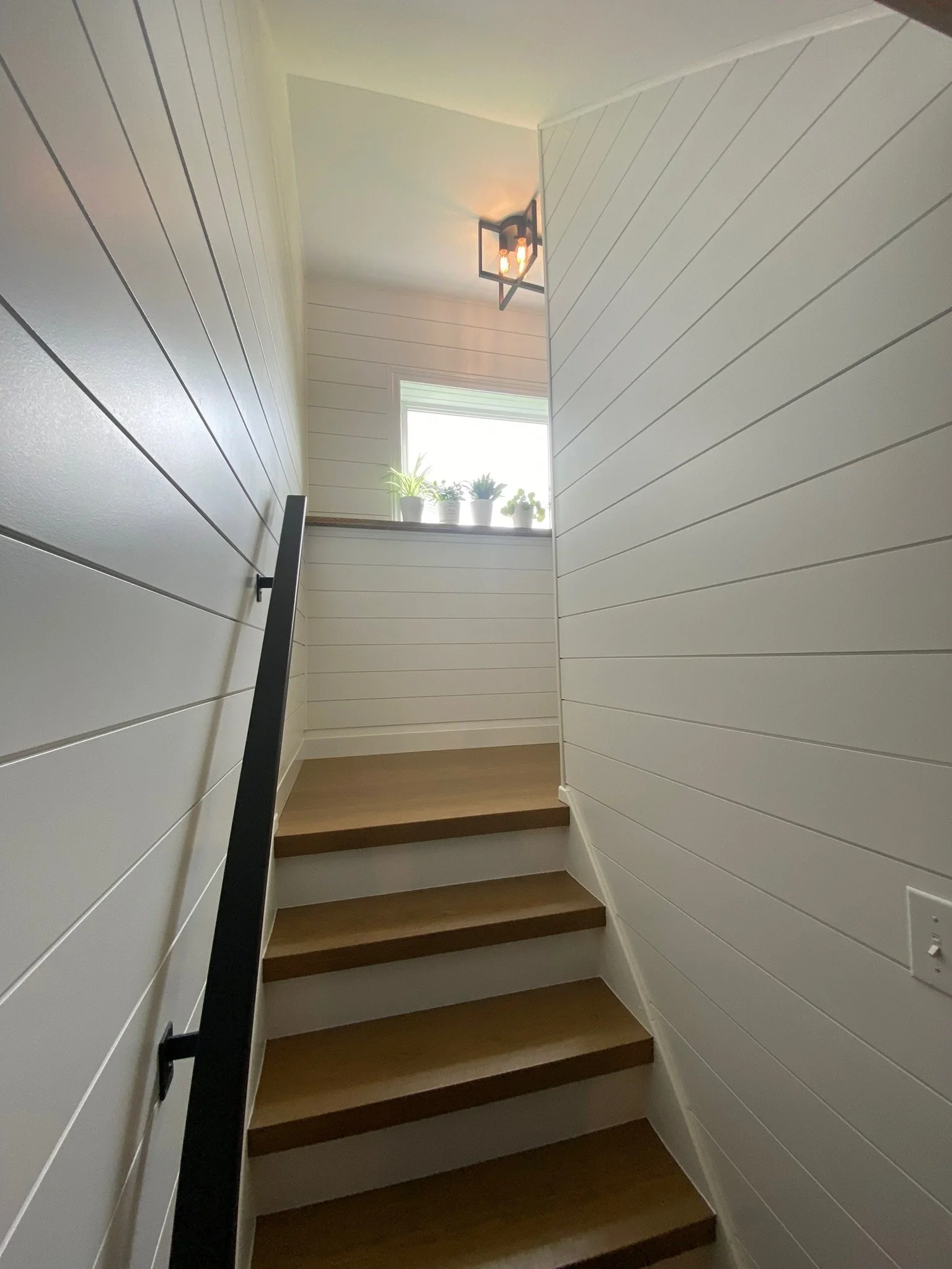View of a wooden staircase with dark steps, white shiplap walls on either side, a black handrail on the left, and a window at the top with potted plants on the windowsill.