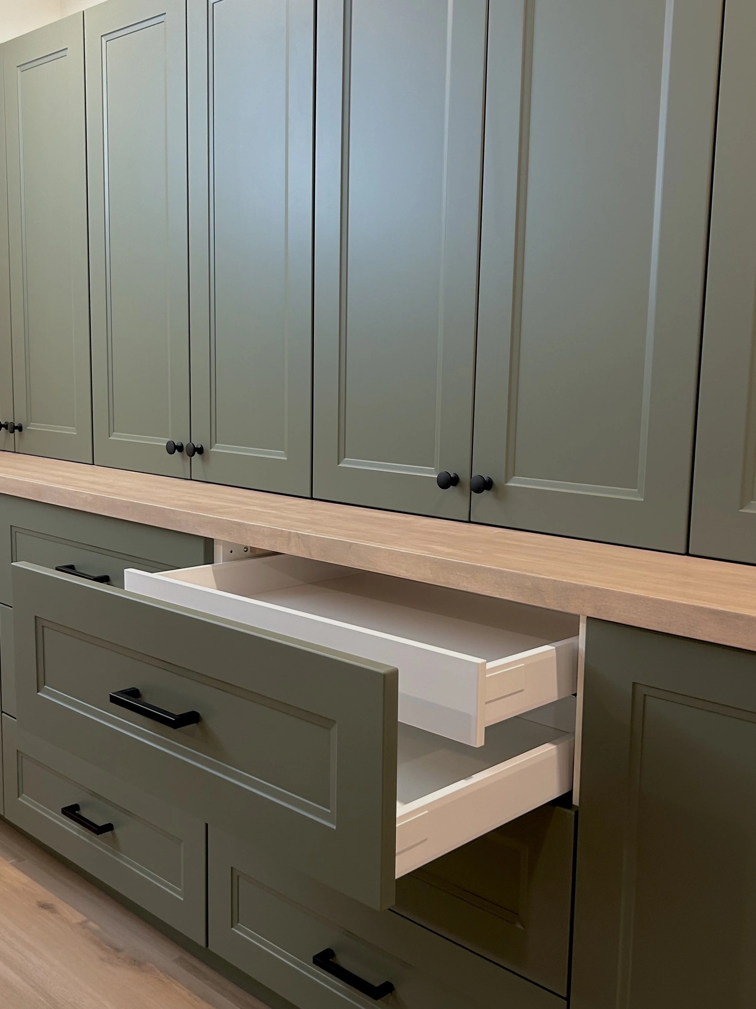 Kitchen cabinets with drawers, one drawer is partially open, painted in a sage green color with black handles, mounted on a wooden countertop, set against a wall.