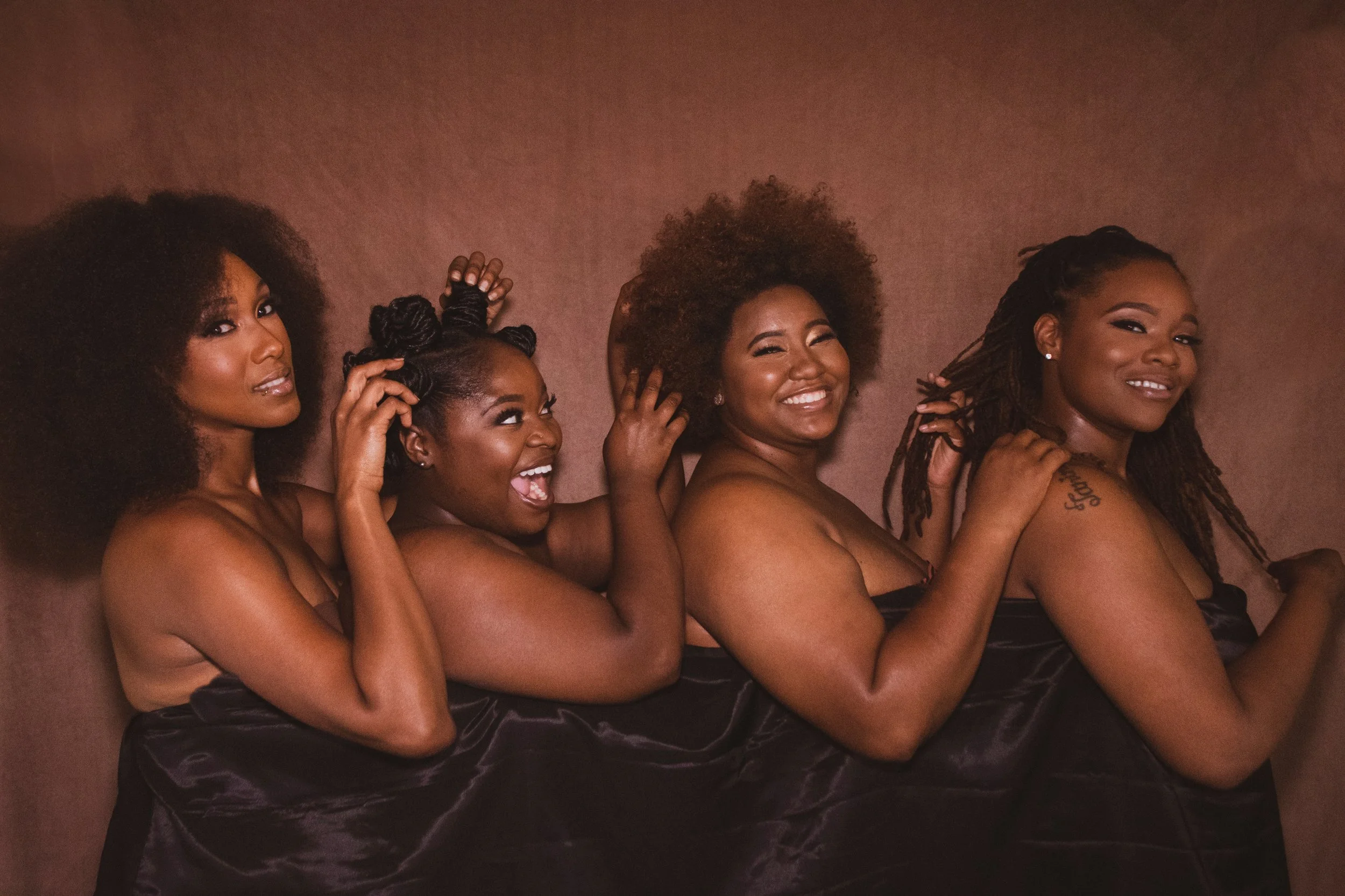 Four women with different hairstyles and smiles, wrapped in black fabric, posing closely together against a brown background.