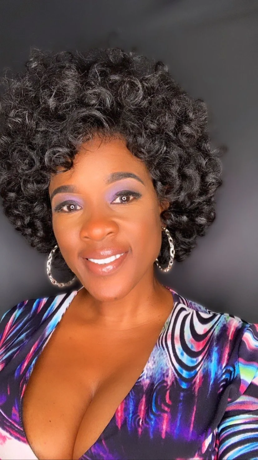 A woman with dark, curly hair and hoop earrings, smiling, wearing a vibrant, patterned top and purple eye makeup against a dark background.