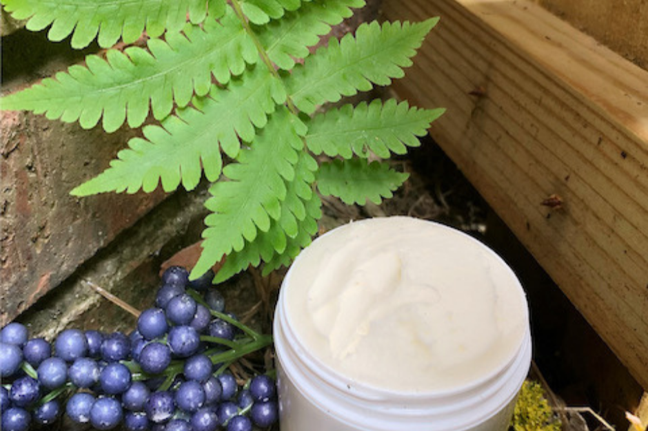 Green fern leaves, dark purple berries, and an open container of cream or lotion on the ground near a brick wall and wooden planks.