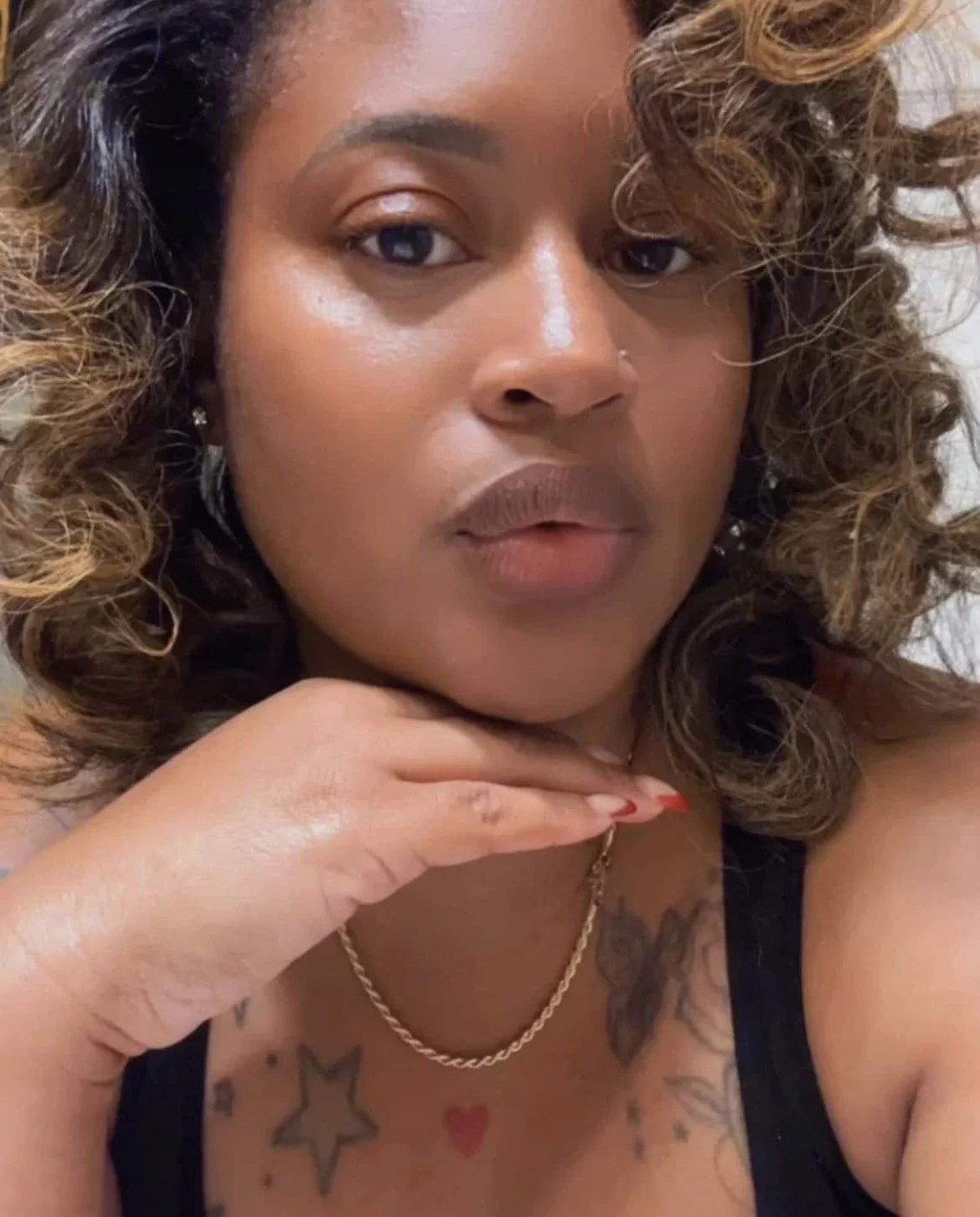 A woman with curly, ombre hair styled in loose ringlets, wearing a black top, a gold chain necklace, and small earrings. Her hand with manicured nails resting near her face, showing tattoos on her chest including a star and a heart.