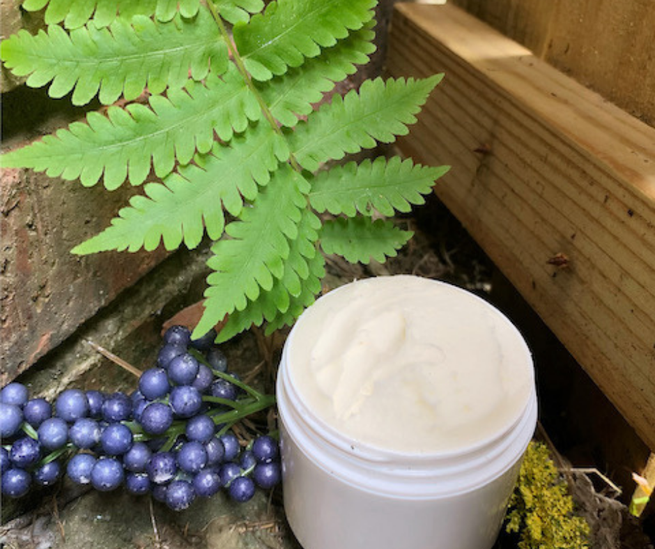 A jar of cream or lotion near green fern leaves, purple berries, and yellow-green moss on the ground.