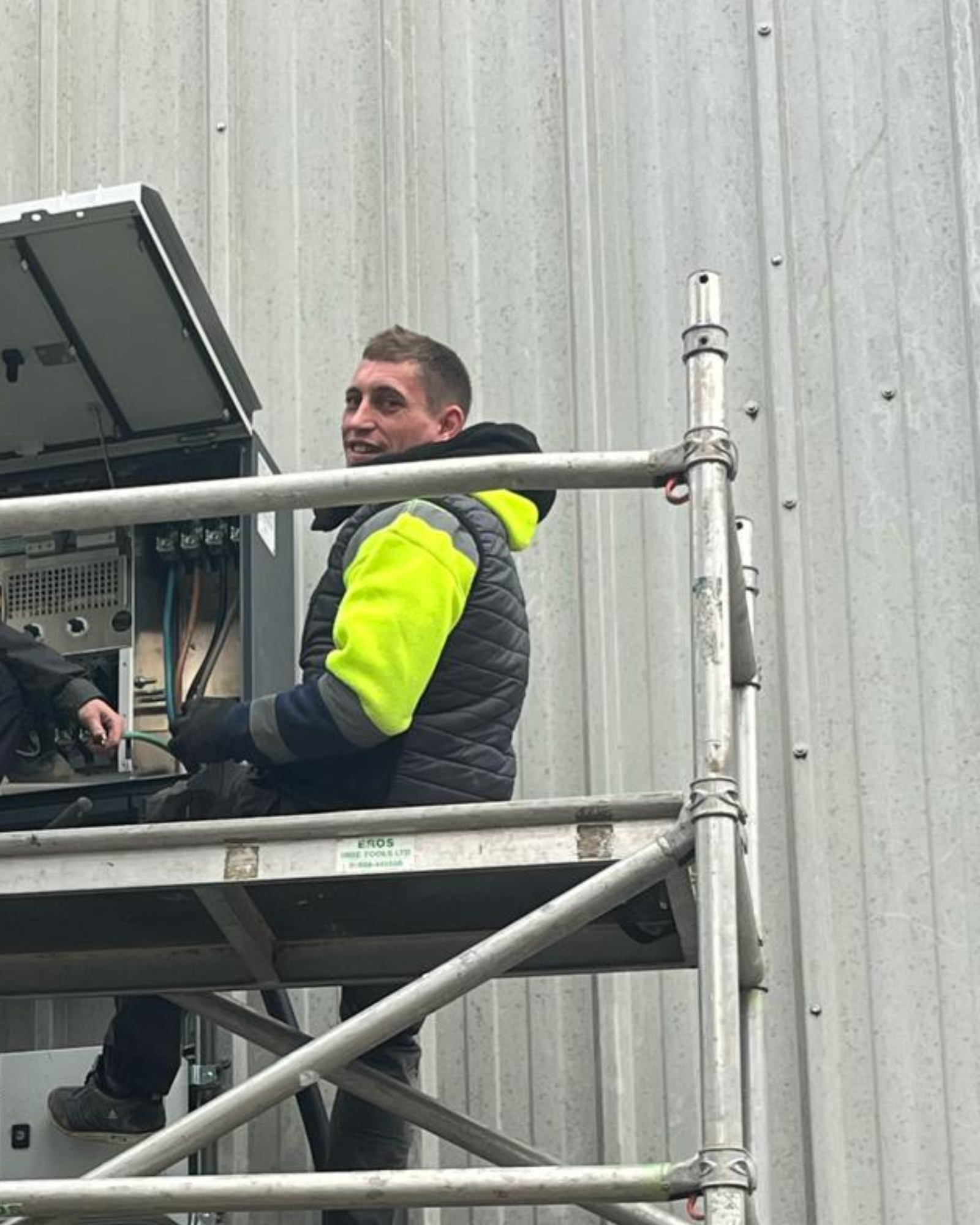 Aaron Boshell, Roof Team Leader in a high-visibility jacket and gloves is on scaffolding, working on an open electrical panel against a corrugated metal wall.