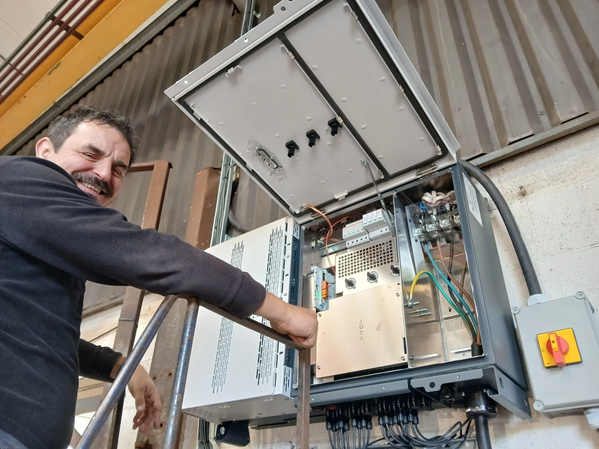 Darren (Daz) Slater, Electrician Technician,  standing beside an open electrical control panel, smiling, with visible wiring and components inside the panel.