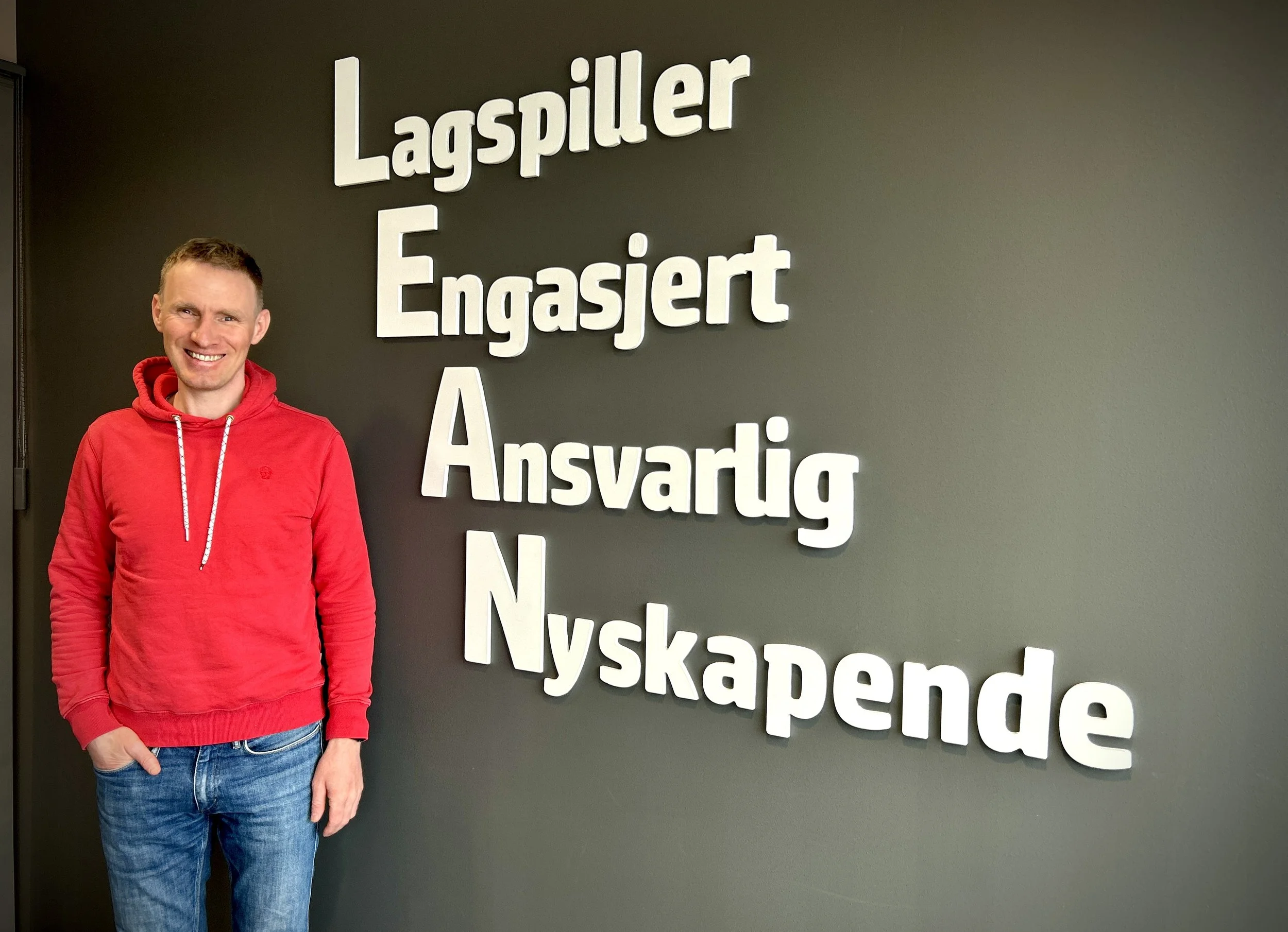 En mann i rød hettegenser står foran en vegg med norske ord som beskriver positive egenskaper, som 'Lagsiller', 'Engasjert', 'Ansvarlig' og 'Nyskapende'.