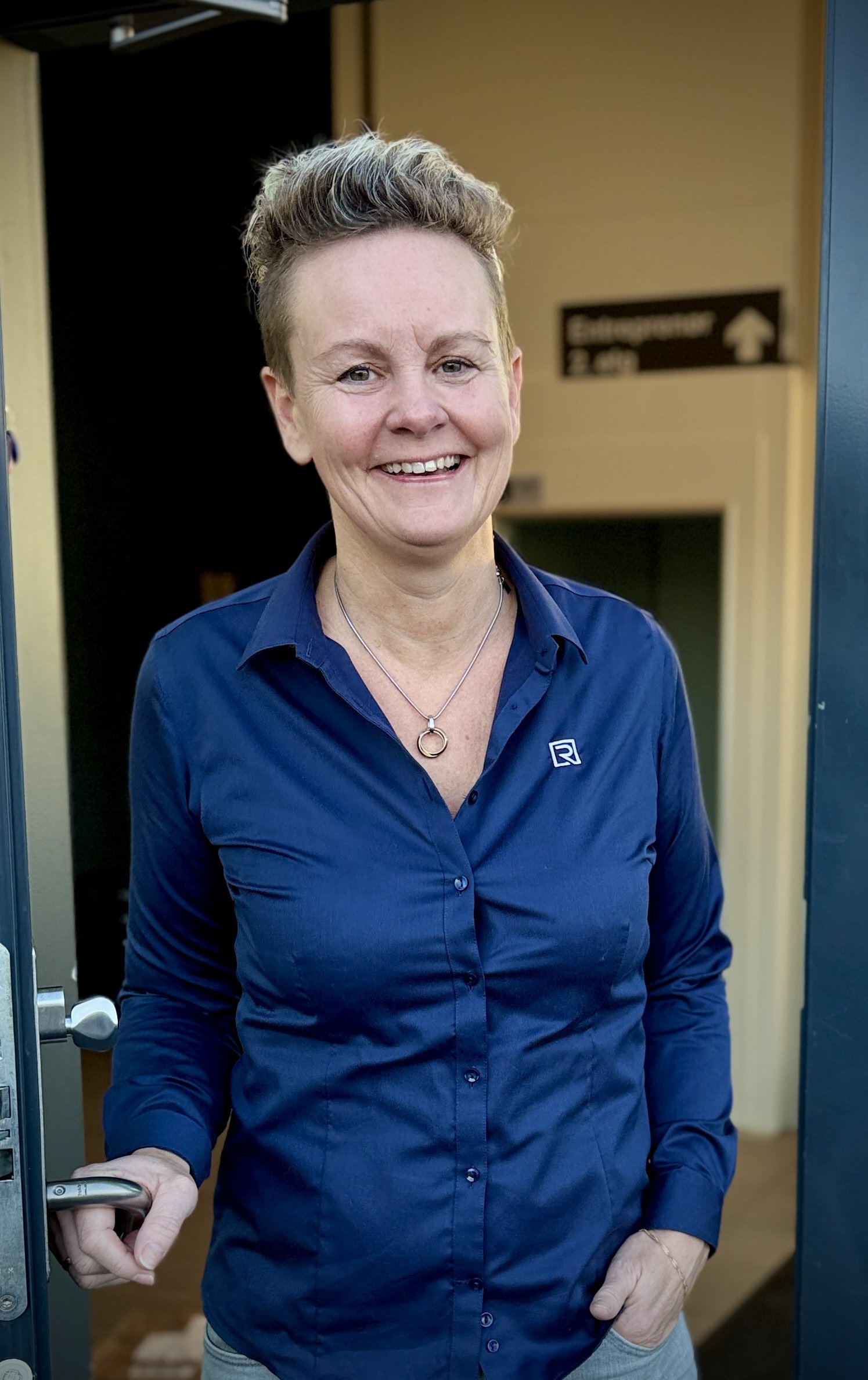 En kvinne med kort, krøllete hår smiler og står i en døråpning, iført en blå knappeskjorte.