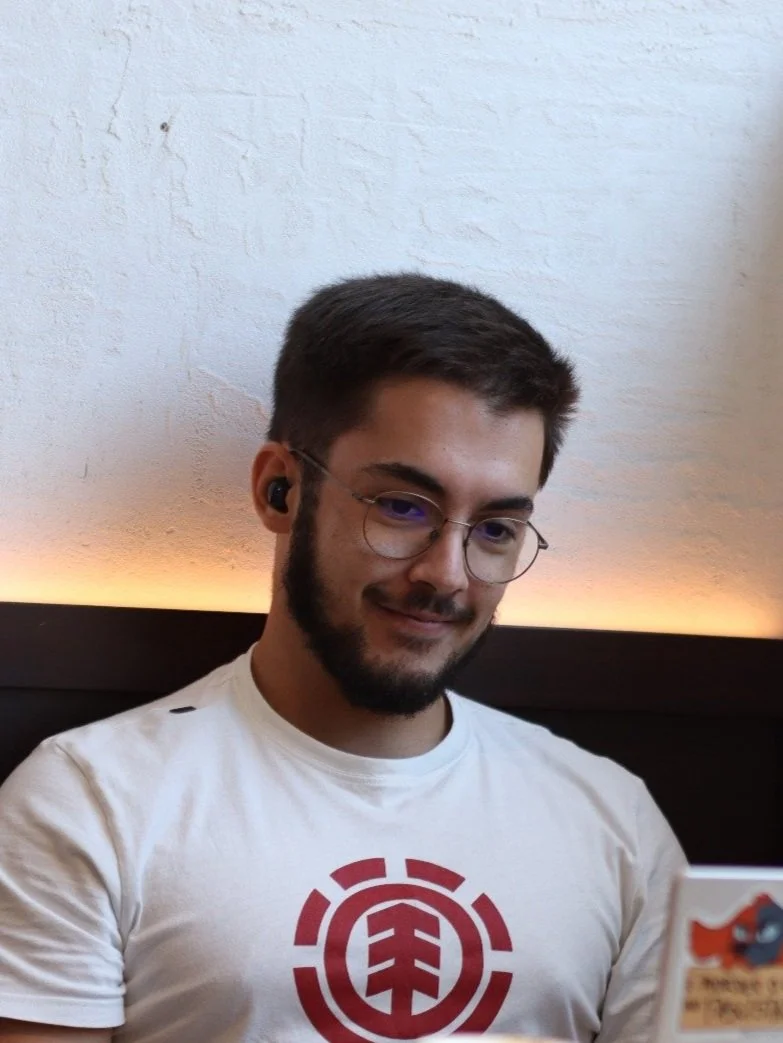 Homem sorrindo usando óculos, camiseta branca com símbolo vermelho, sentado contra uma parede branca com iluminação quente ao fundo.