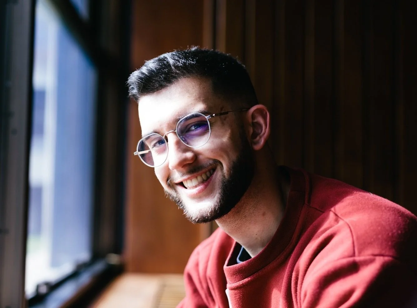 Jovem sorridente com óculos de armação redonda, usando suéter vermelho, sentado perto de uma janela com fundo de painéis de madeira.
