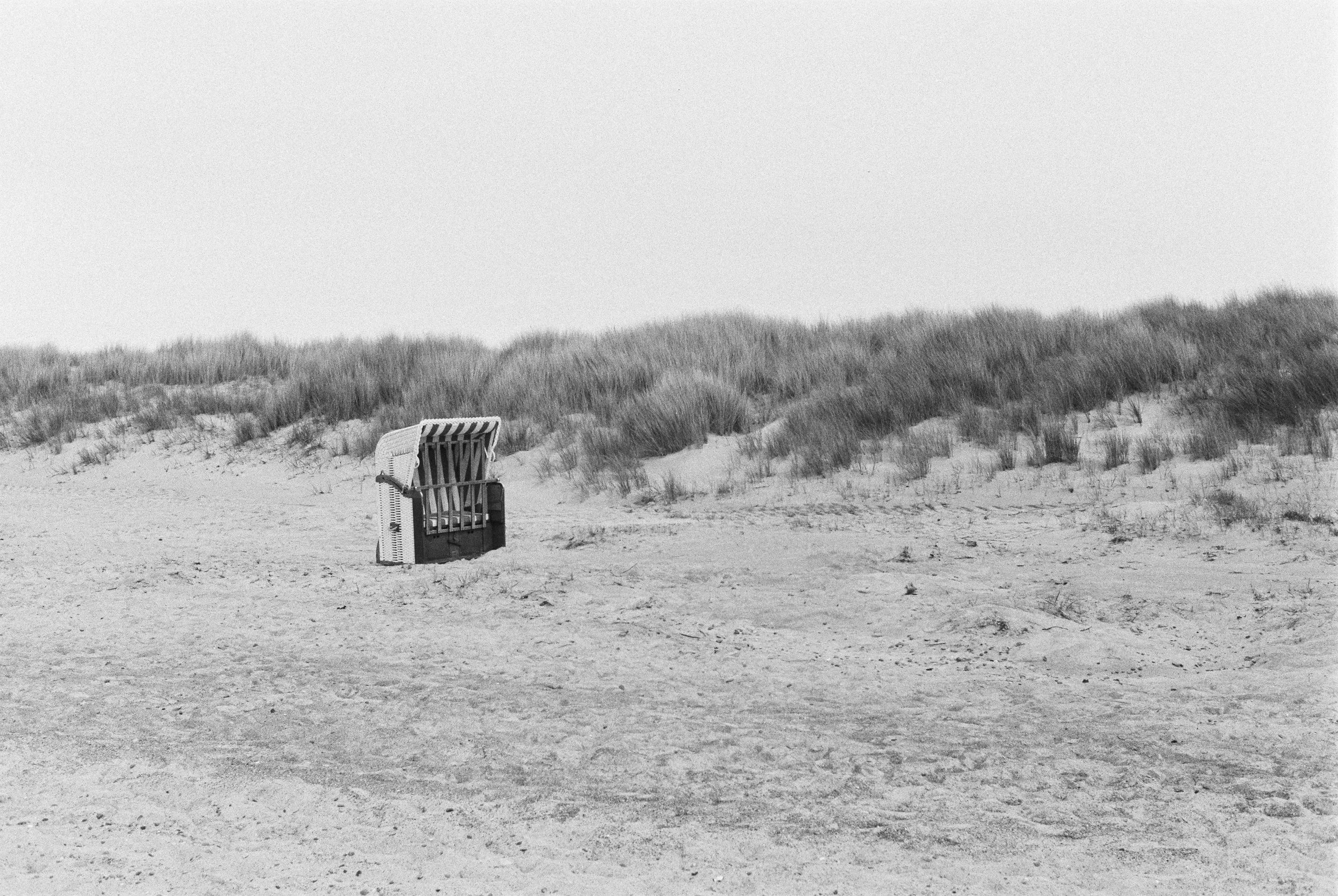 Ein einzelner Strandkorb auf einem verlassenen Sandstrand mit Dünen im Hintergrund, in Schwarz-Weiß, keine Menschen sichtbar. Diernhagen auf BW-Film.