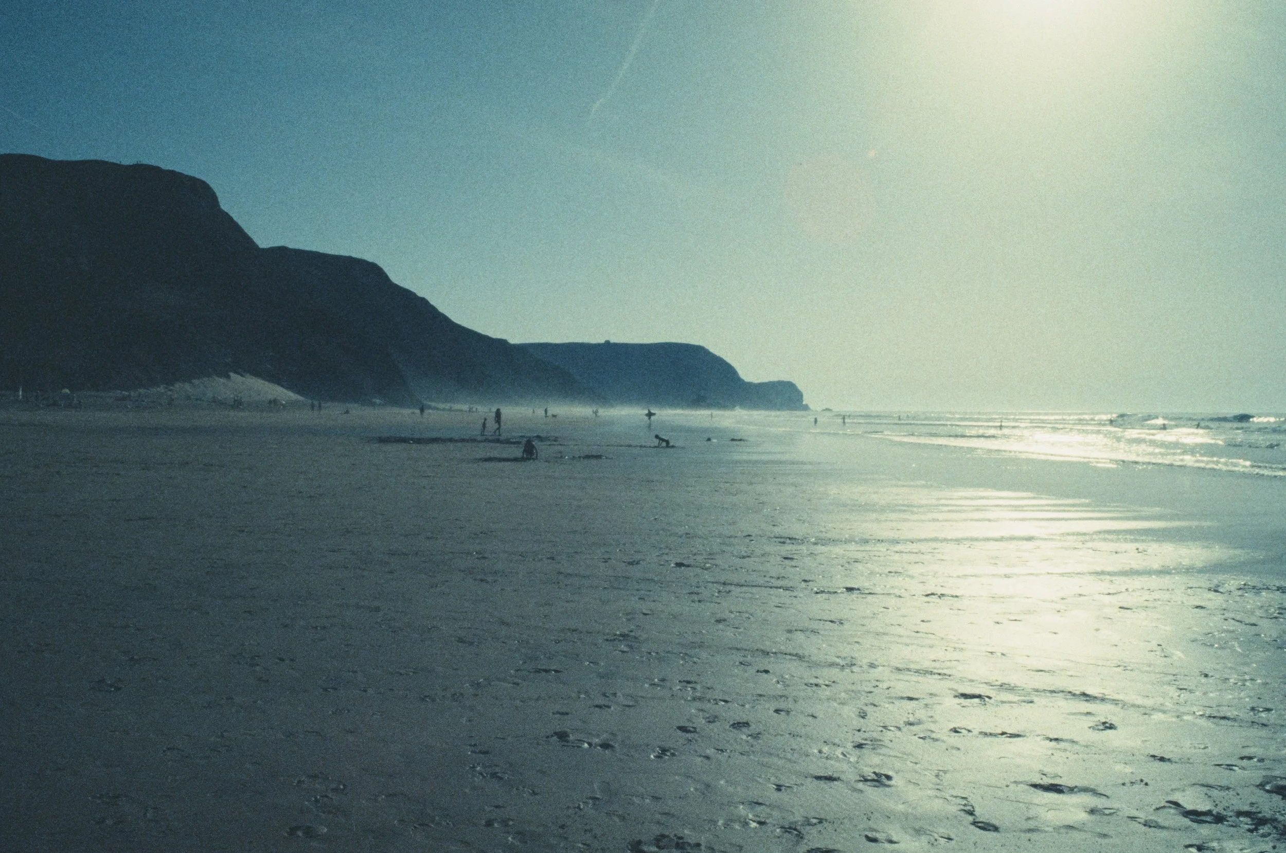 Strand mit Menschen, die am Wasser spazieren gehen, große Klippen im Hintergrund, Sonnenlicht spiegelt sich auf dem Wasser. Retrochrome Diafilm.