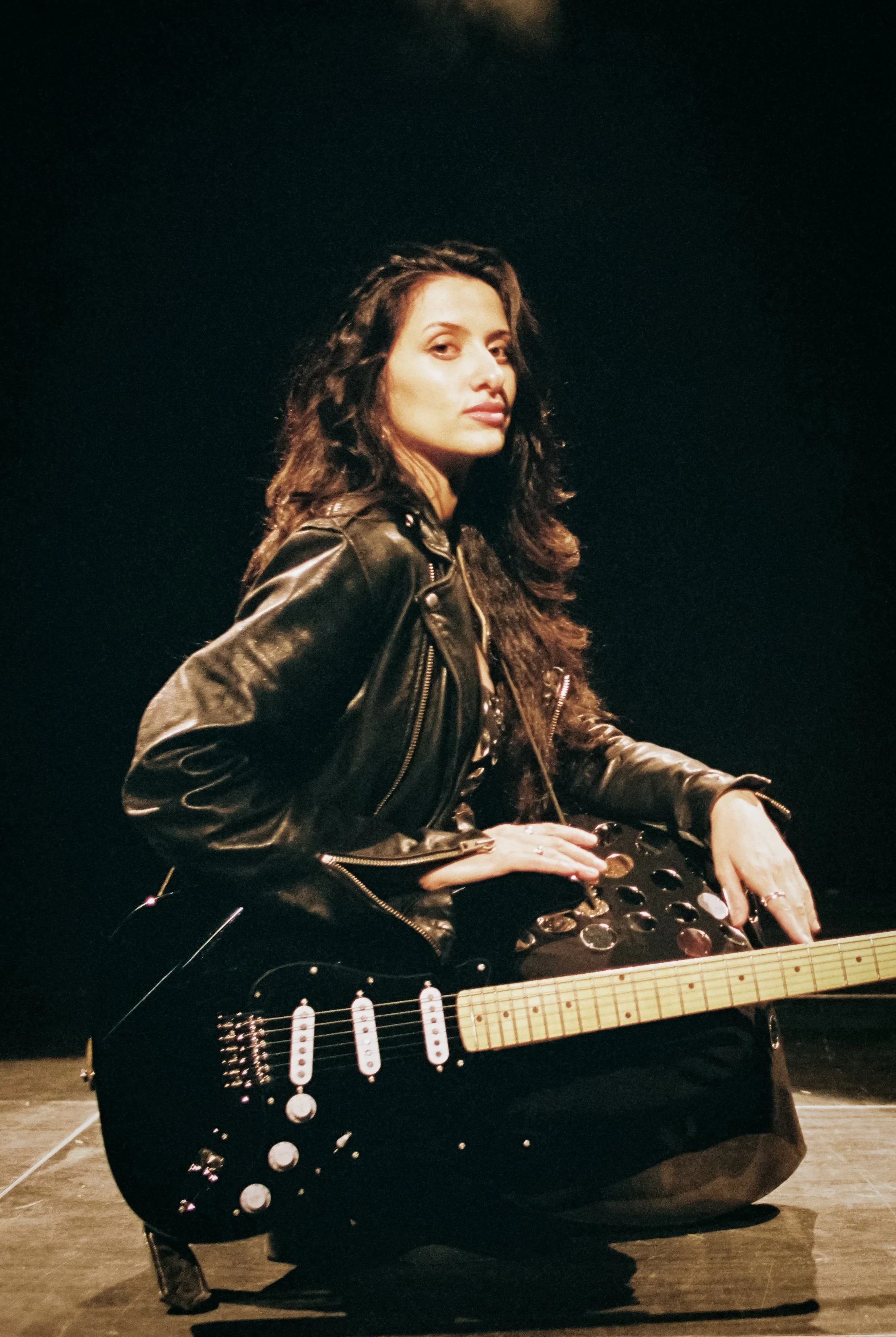Elif Batman bekannt aus The Voice mit dunklen, lockigen Haaren spielt eine schwarze Harley Benton St-57 E-Gitarre auf einer Bühne, trägt eine schwarze Lederjacke und sitzt auf dem Boden vor einem schwarzen Hintergrund. Geschossen auf Portra 400.