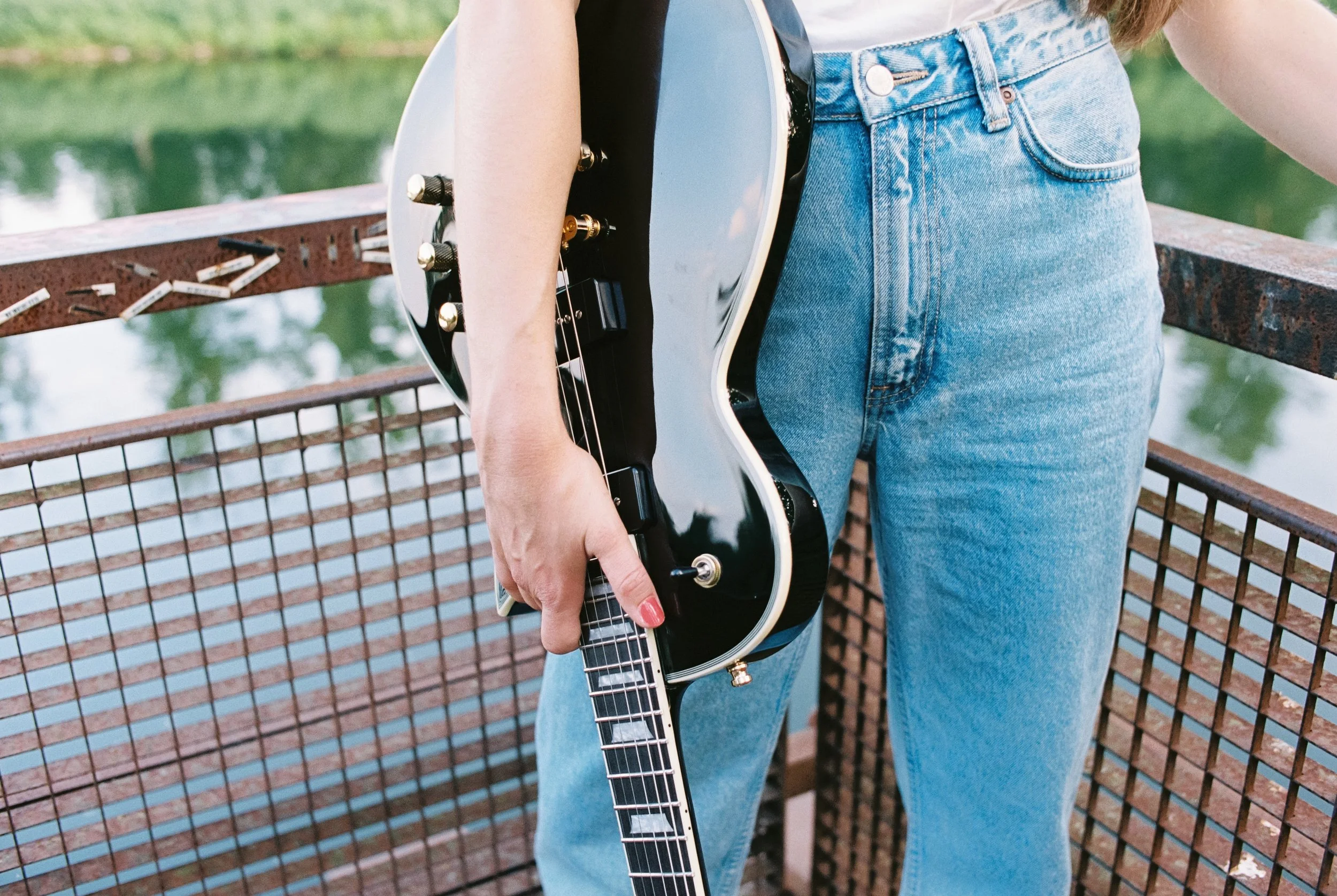 Nahaufnahme einer Person in Blue Jeans, die eine schwarze Harley Bneton SC-Custom P90 Gitarre hält, während sie auf einer Brücke steht. Geschossen auf Portra 400 mit der Nikon F2.