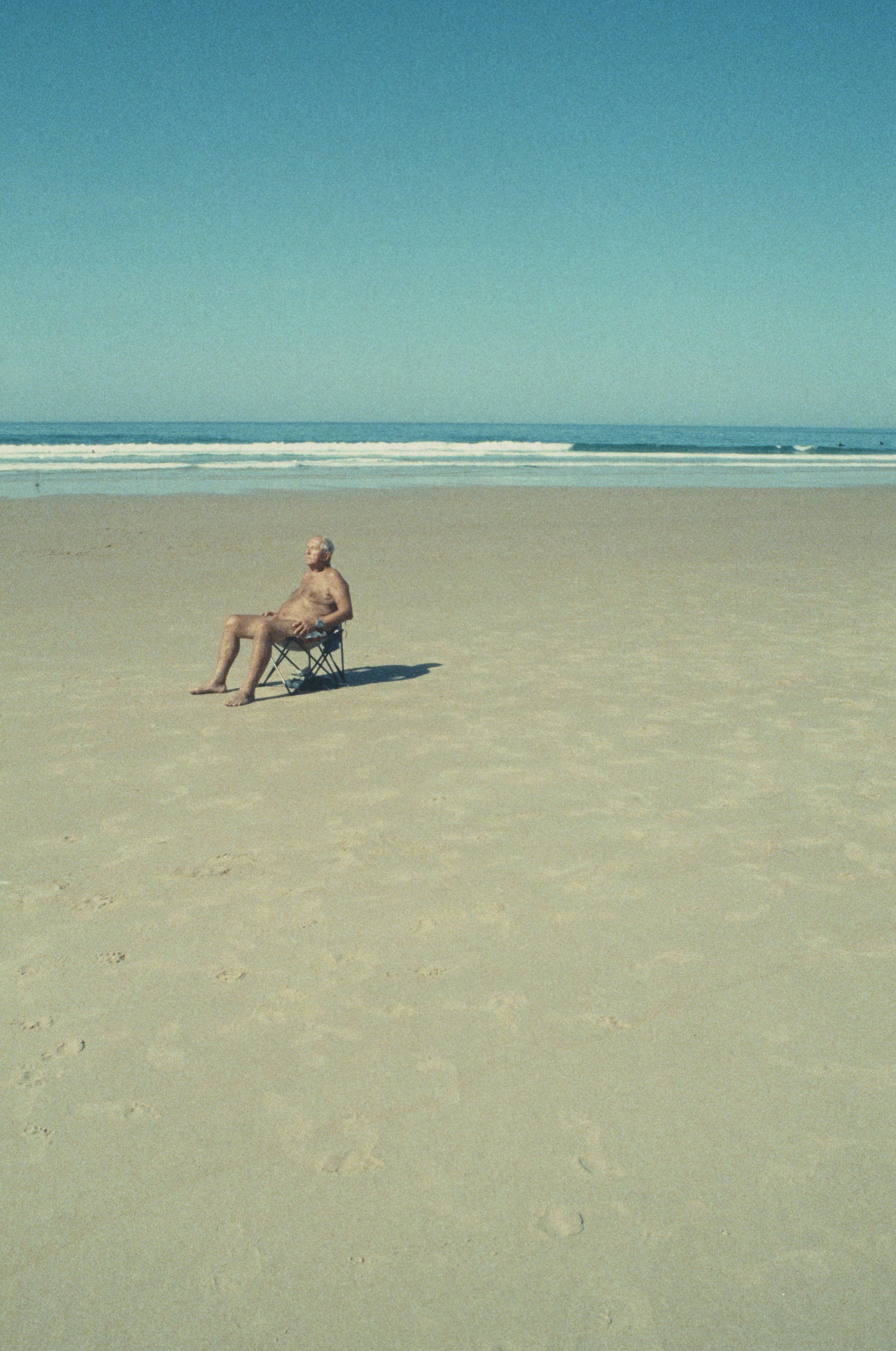 Ein älterer Mann sitzt entspannt in einem Klappstuhl auf einem einsamen Strand, der Sand ist hell und der Himmel klar. Das Meer ist im Hintergrund mit Wellen. Fotografiert auf Retrochrom Diafilm.