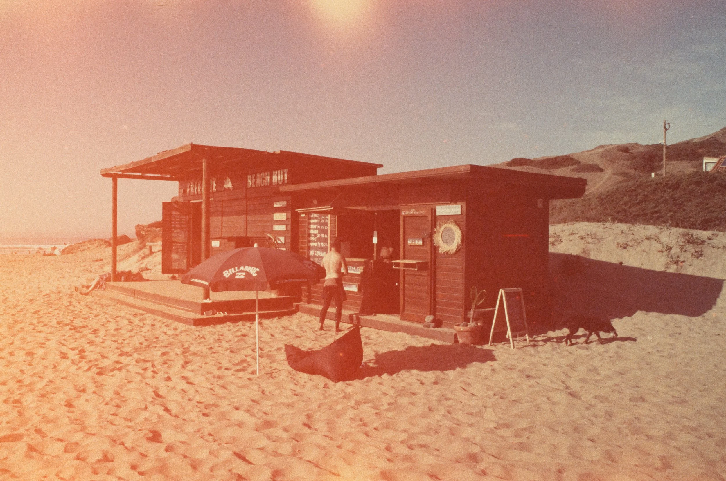 Ein Strandcafé aus Holz mit einem Sonnenschirm, Sand und einigen Personen und Dächern in den Dünen im Hintergrund. Fotografiert auf abgelaufenem Diafilm.