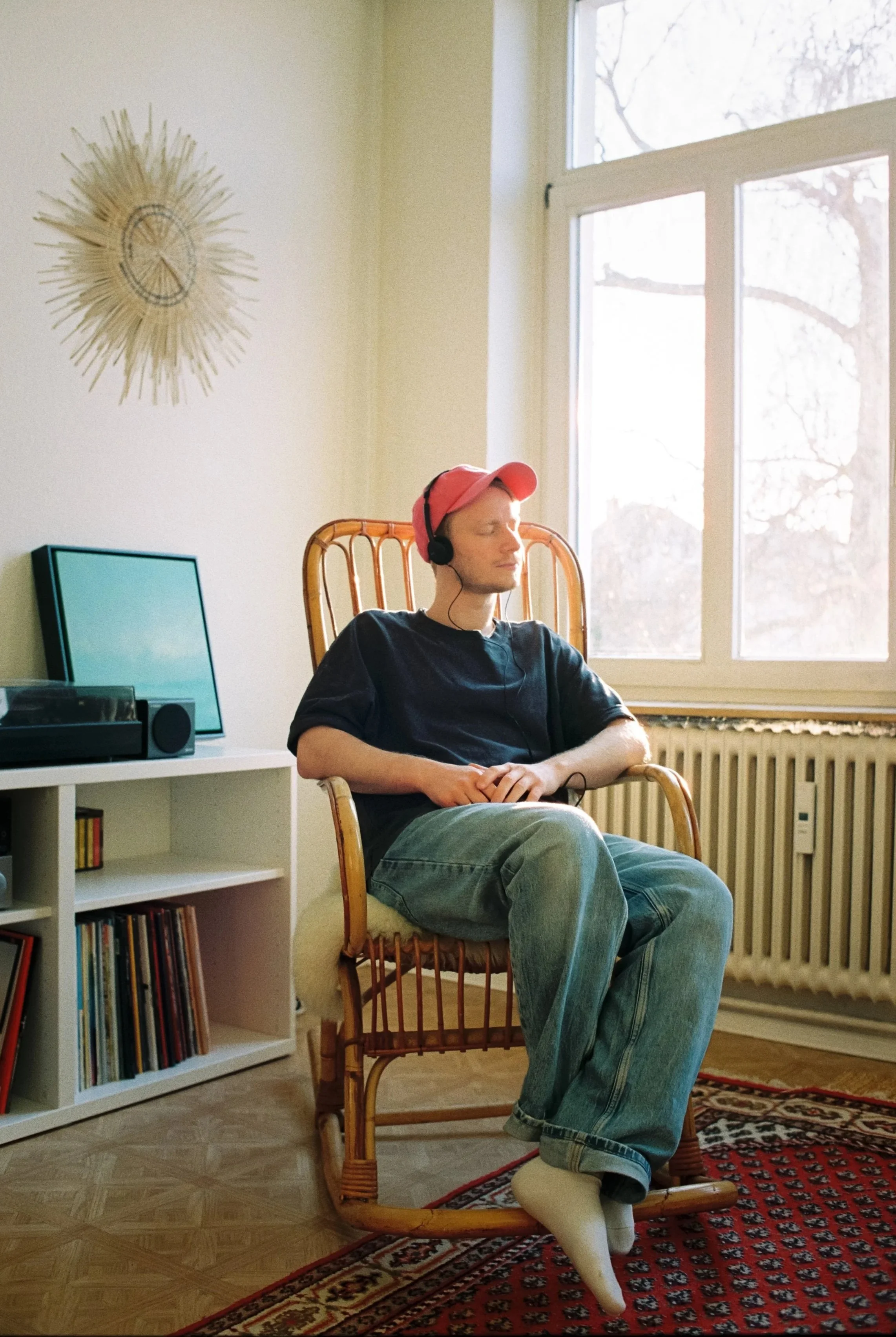 Junge Person sitzt entspannt in einem Rattanstuhl mit geschlossenen Augen, trägt Kopfhörer, eine rote Kappe, schwarze T-Shirt, Jeans, und trägt weiße Socken, vor einem Fenster mit Blick nach draußen in einem gemütlichen Raum mit einem Regal voller Schallplatten.