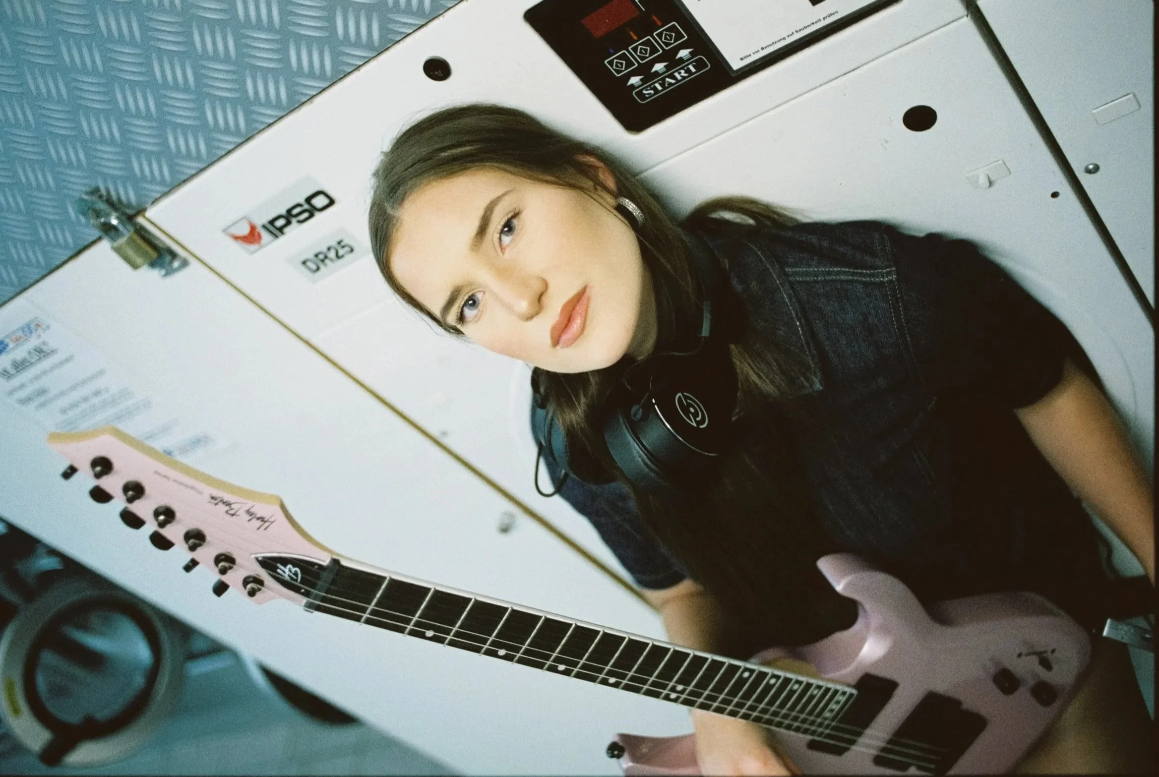 Junge Frau mit einem pinken E-Gitarren, liegend in einer Waschmaschine, trägt schwarze Jacke und Kopfhörer, im Hintergrund eine Waschmaschine und eine blaue Wand.
