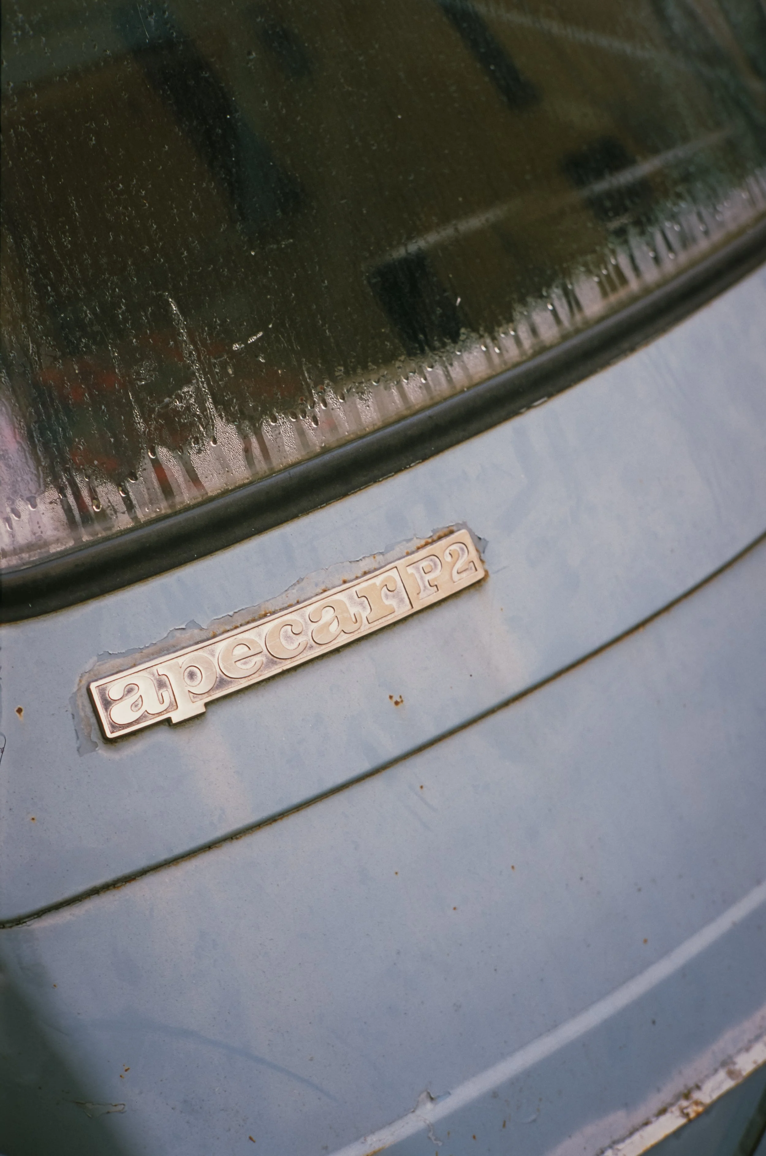Nahaufnahme eines blauen Autos mit einem alten, rostigen Emblem, das den Namen 'apecarr P2' trägt, auf der Motorhaube. Im Hintergrund sind Fenster und Regentropfen auf der Windschutzscheibe sichtbar. Orte VT auf Ektachrome.
