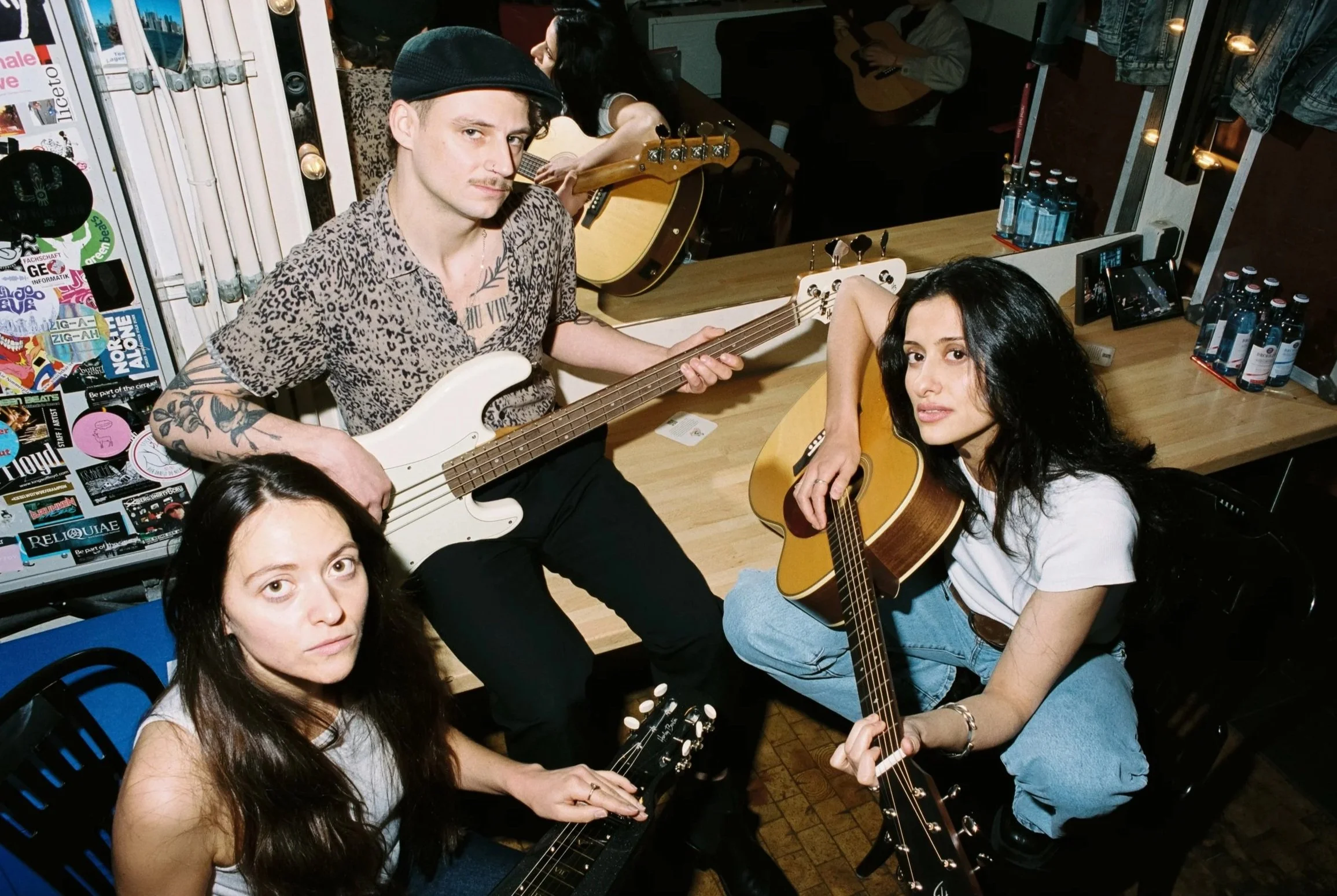 Vier Menschen mit Gitarren in einem Raum, die sich auf eine musikalische Session vorbereiten, mit einem Spiegel im Hintergrund und Getränken auf einem Tisch.