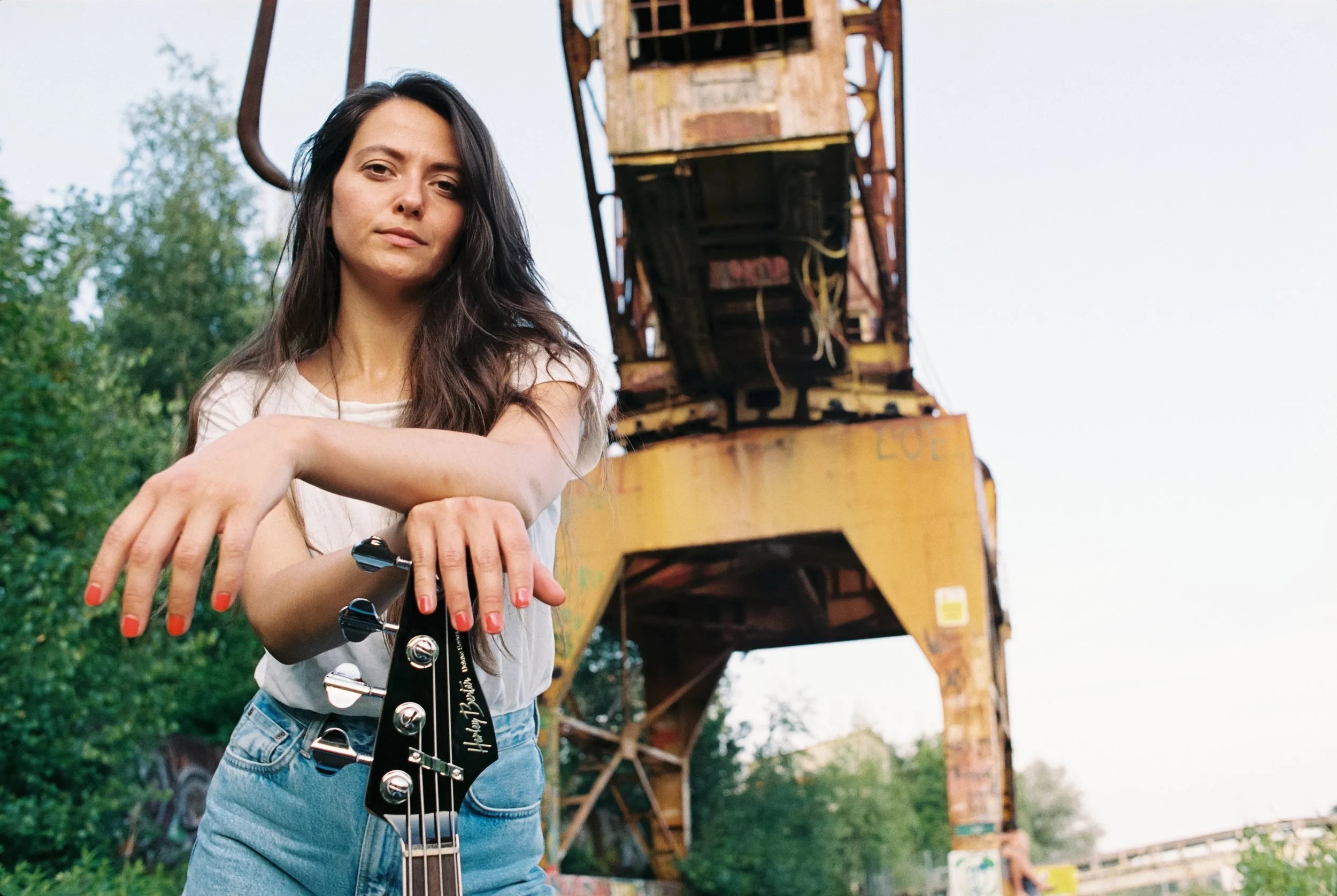 Junge Frau mit dunklen Haaren, die eine E-Gitarre hält, vor einem alten, gelben Kran im Freien, umgeben von Bäumen.