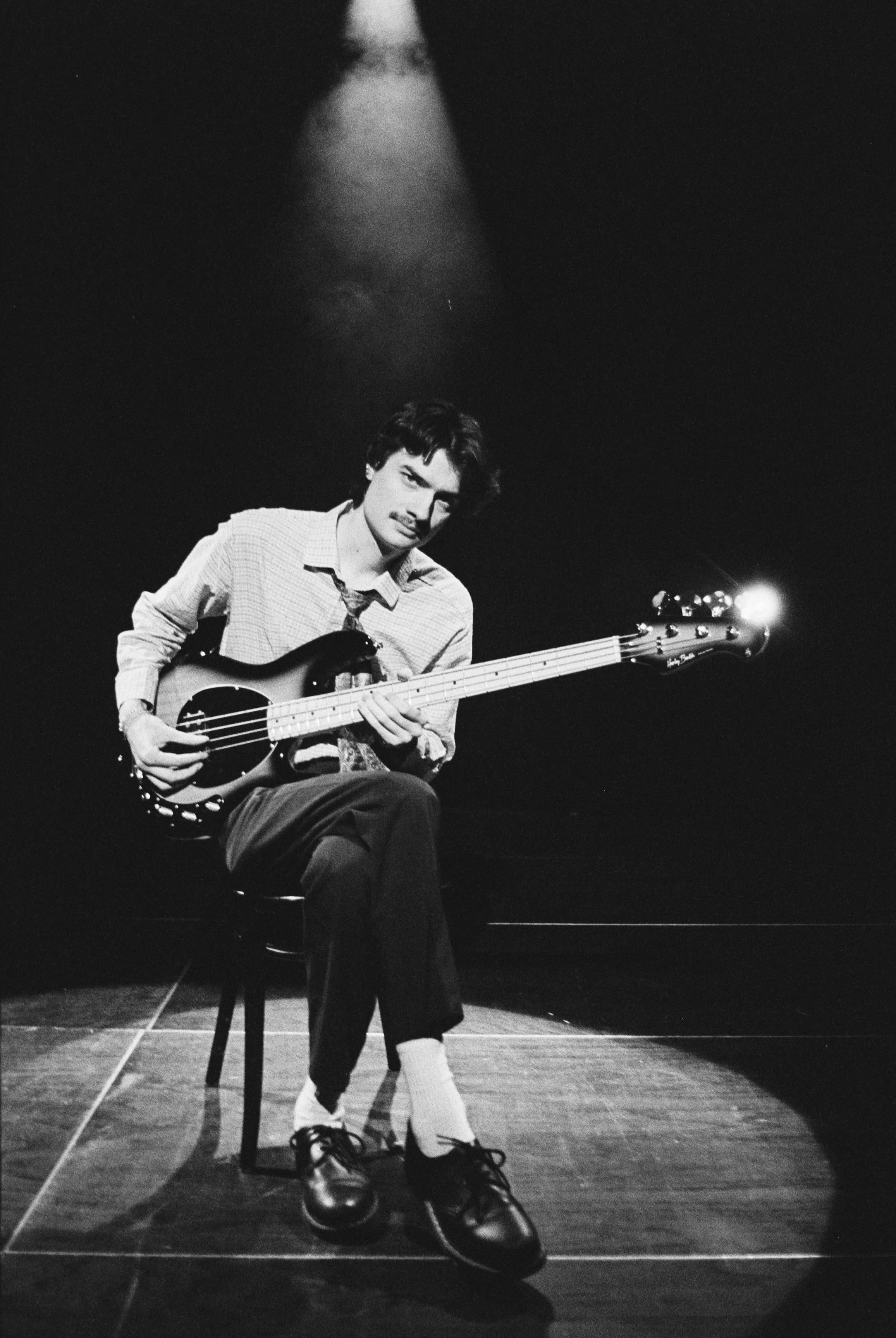Schwarzes und weißes Foto eines jungen Mannes, der auf einem Stuhl sitzt, ein E-Bass spielt und unter einem Spotlicht steht.