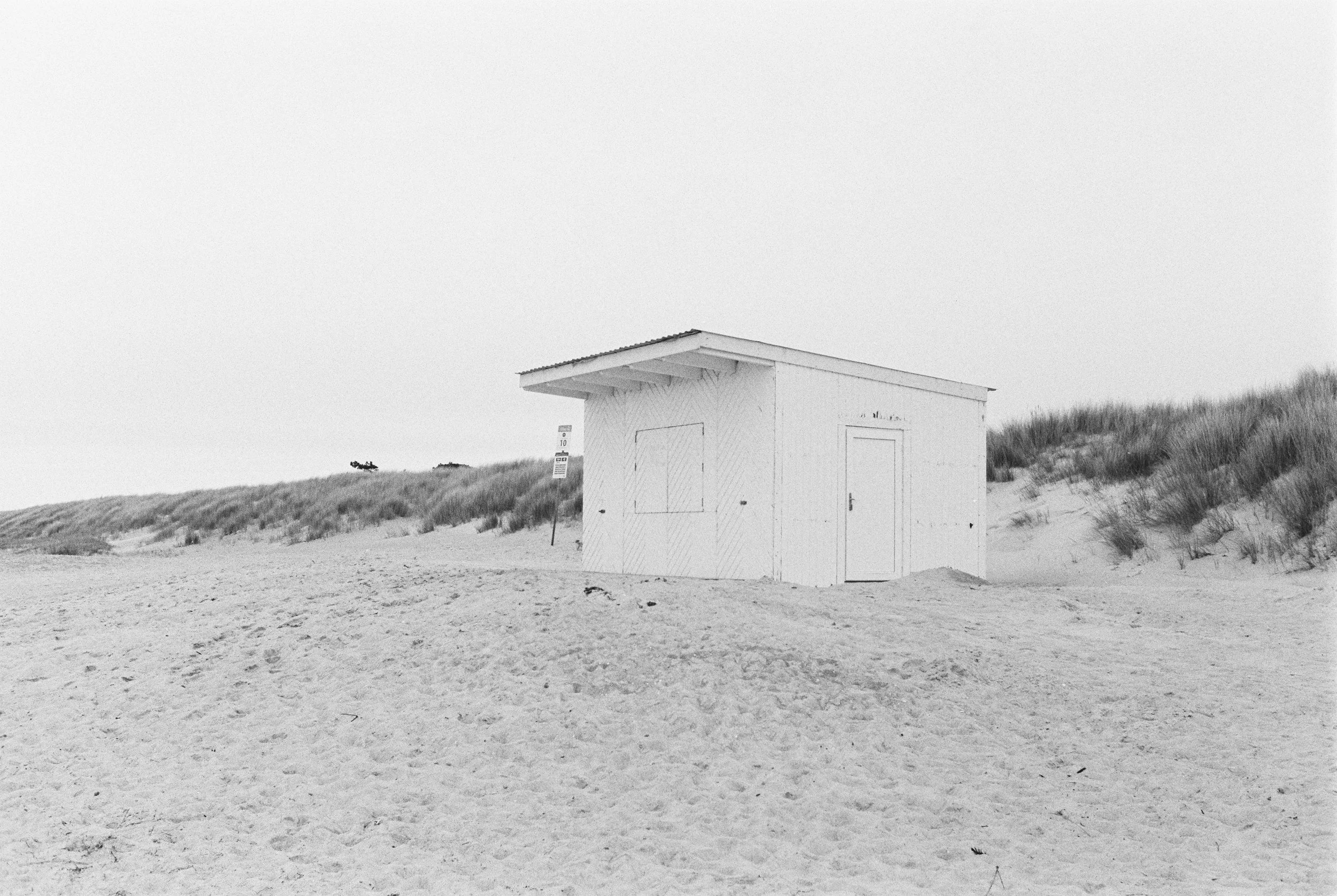 Ein weißes Strandhäuschen auf einem Sandstrand mit Dünen im Hintergrund, bei hellem Himmel. Diernhagen auf BW-Film.