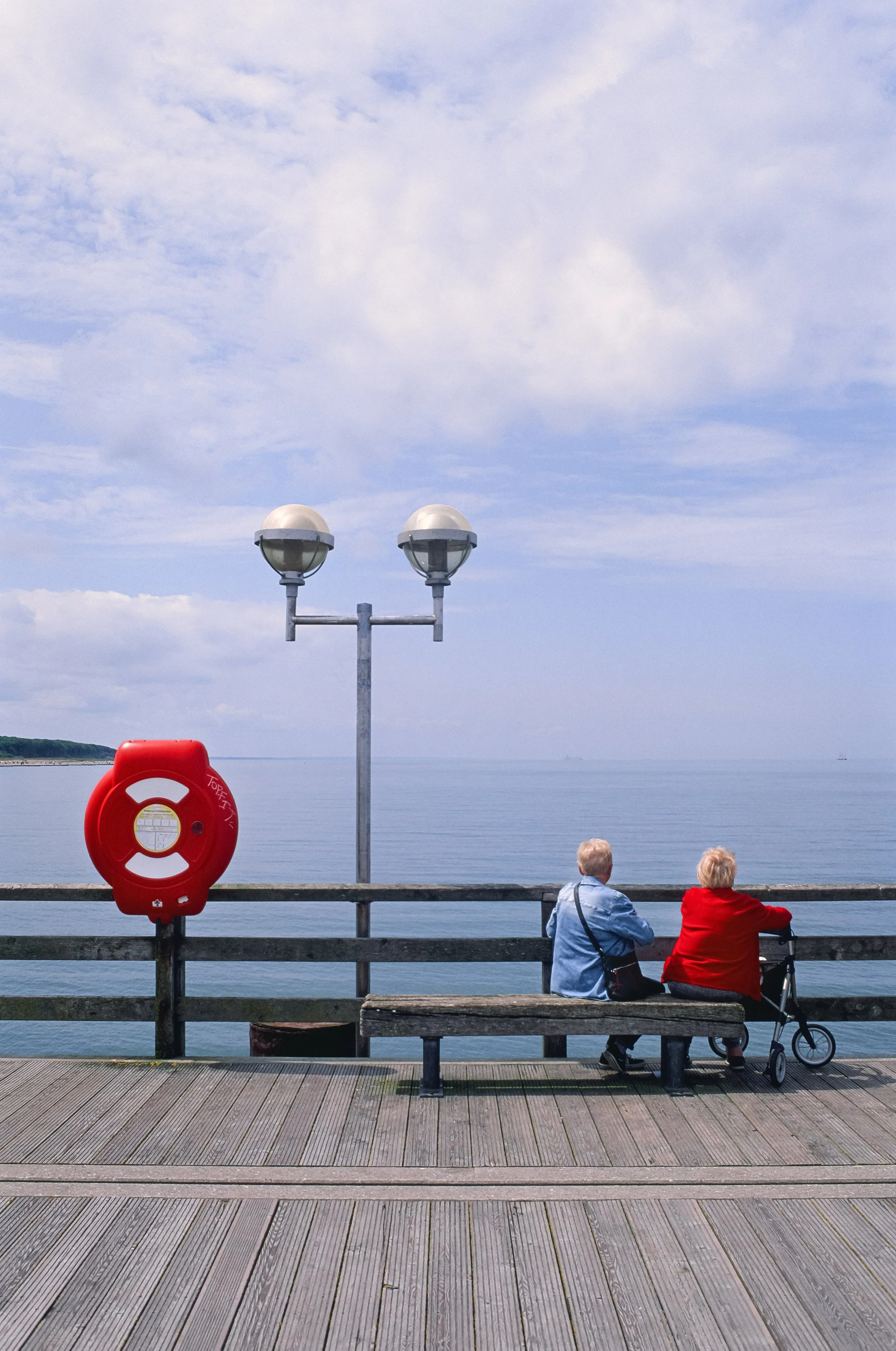 Zwei ältere Frauen sitzen an einem Hafenaufbau aus Holz, mit Blick aufs Meer, neben einem Rollator und einer Rettungsleine, im Hintergrund Himmel und Wasser. Diernhagen strand auf Fuji Velvia.